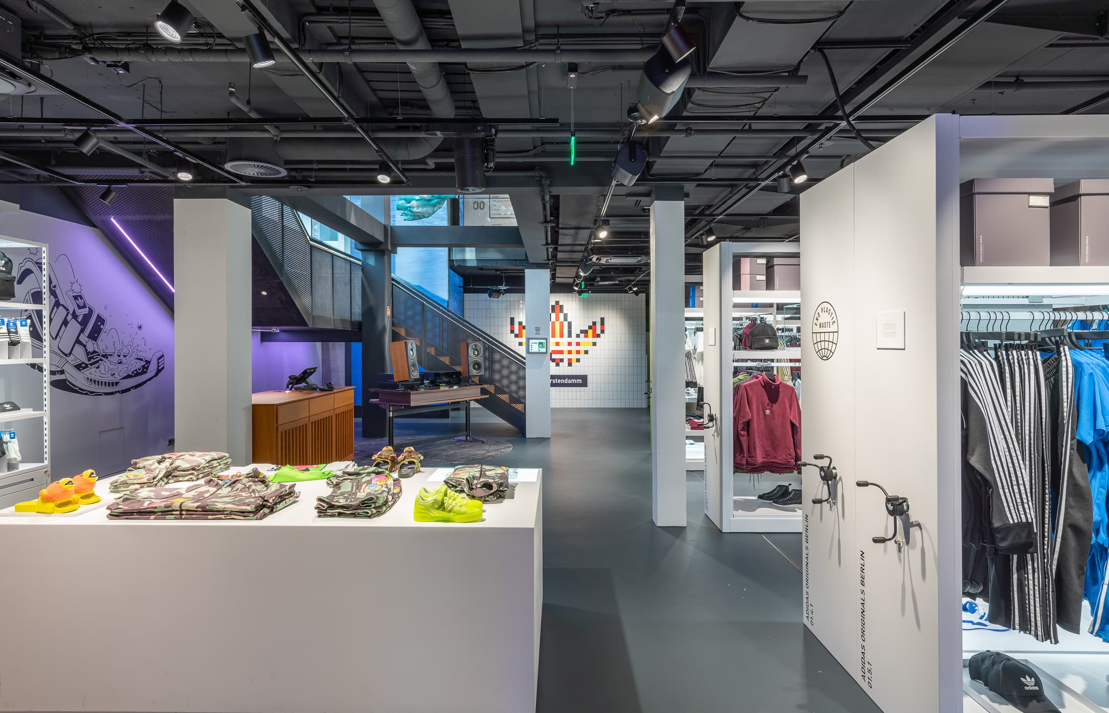 Sportgeschäft mit Bekleidungsständern, Schuhen auf Präsentationstischen und offen sichtbarer Deckentechnik.
