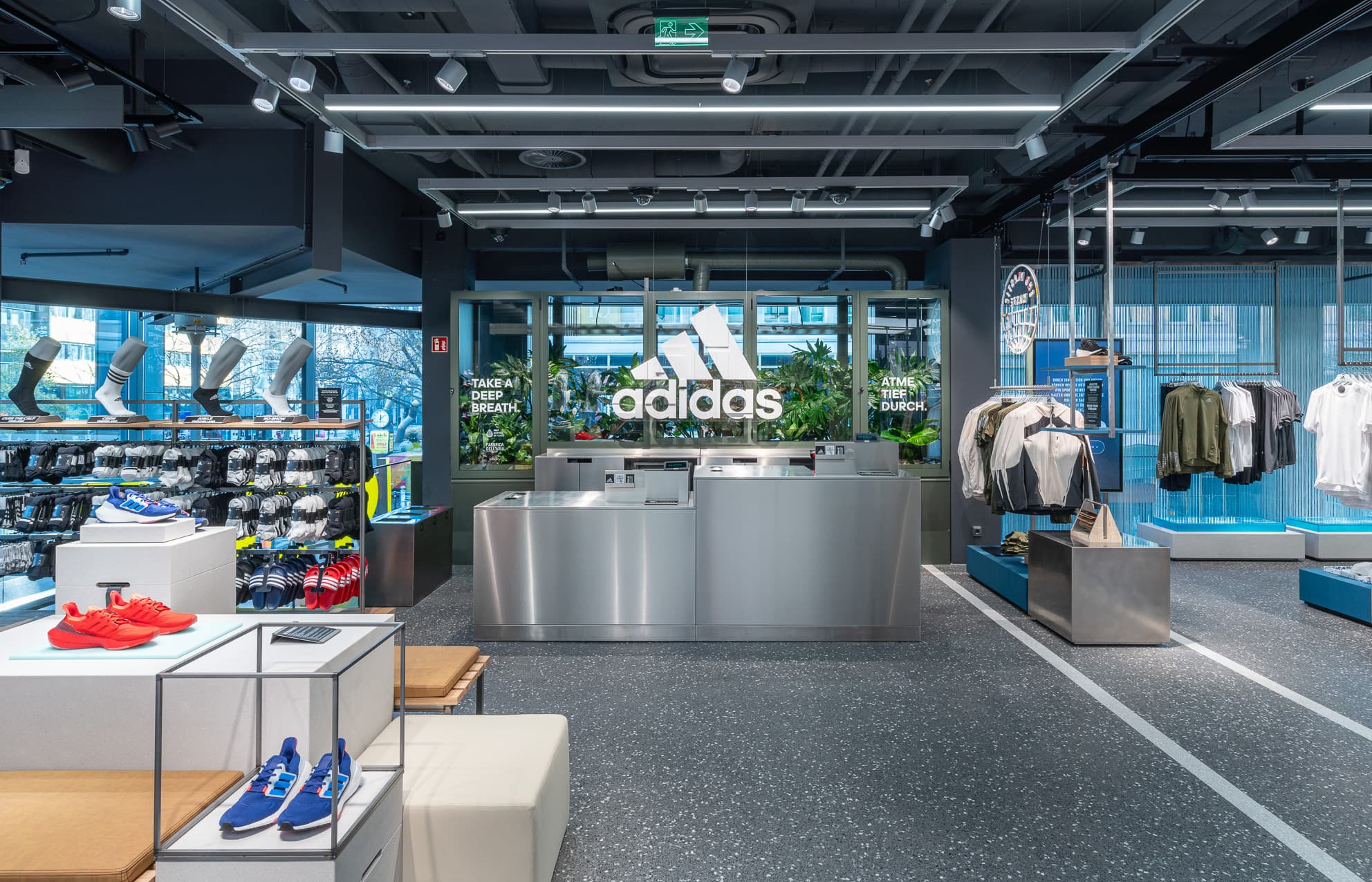 Adidas-Sportgeschäft mit Kassentresen, Schuhpräsentationen, Bekleidungsständern und begrüntem Markenlogo.