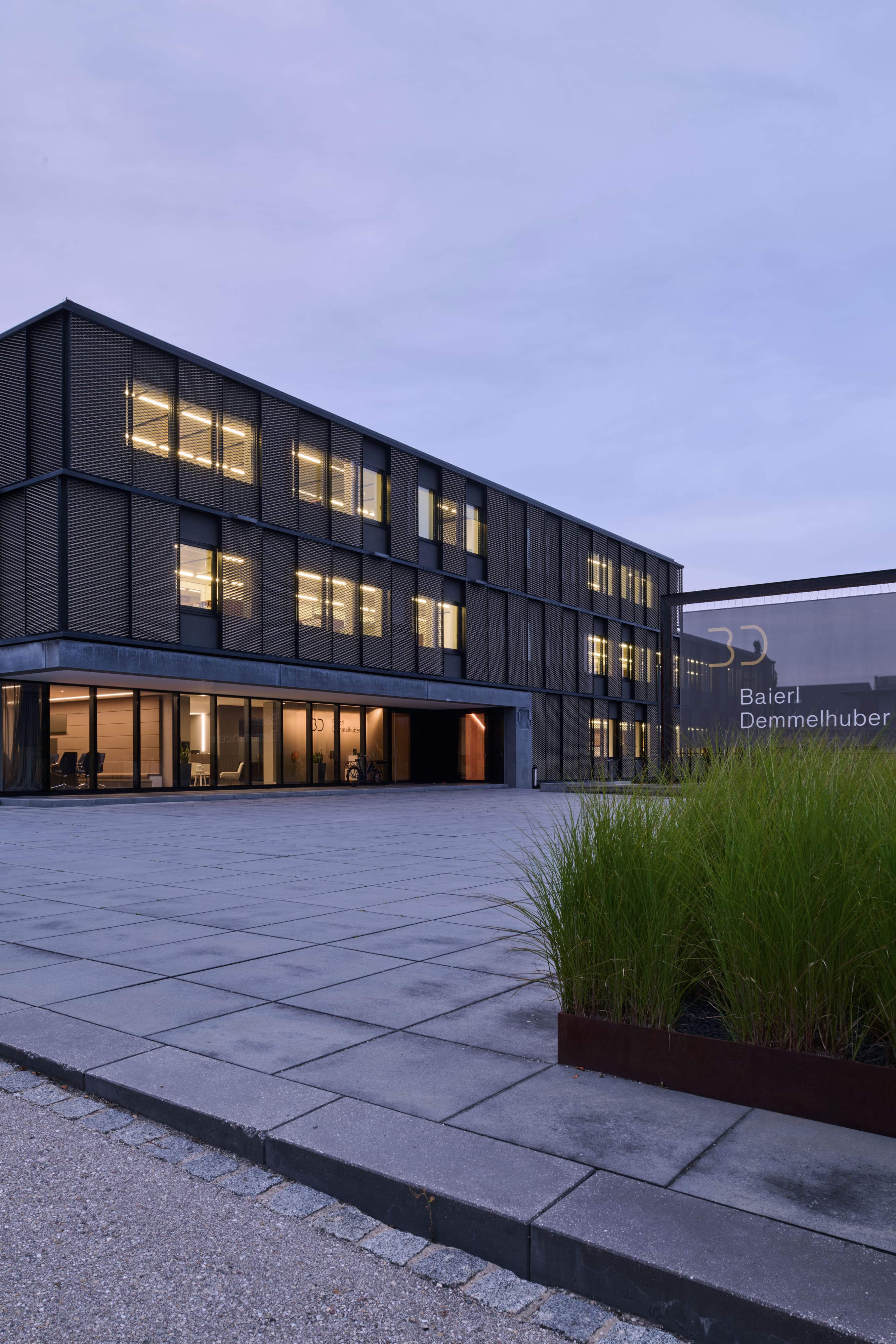 Modernes Firmengebäude mit dunkler Fassade, Vorplatz und begrünten Außenanlagen in der Abenddämmerung.