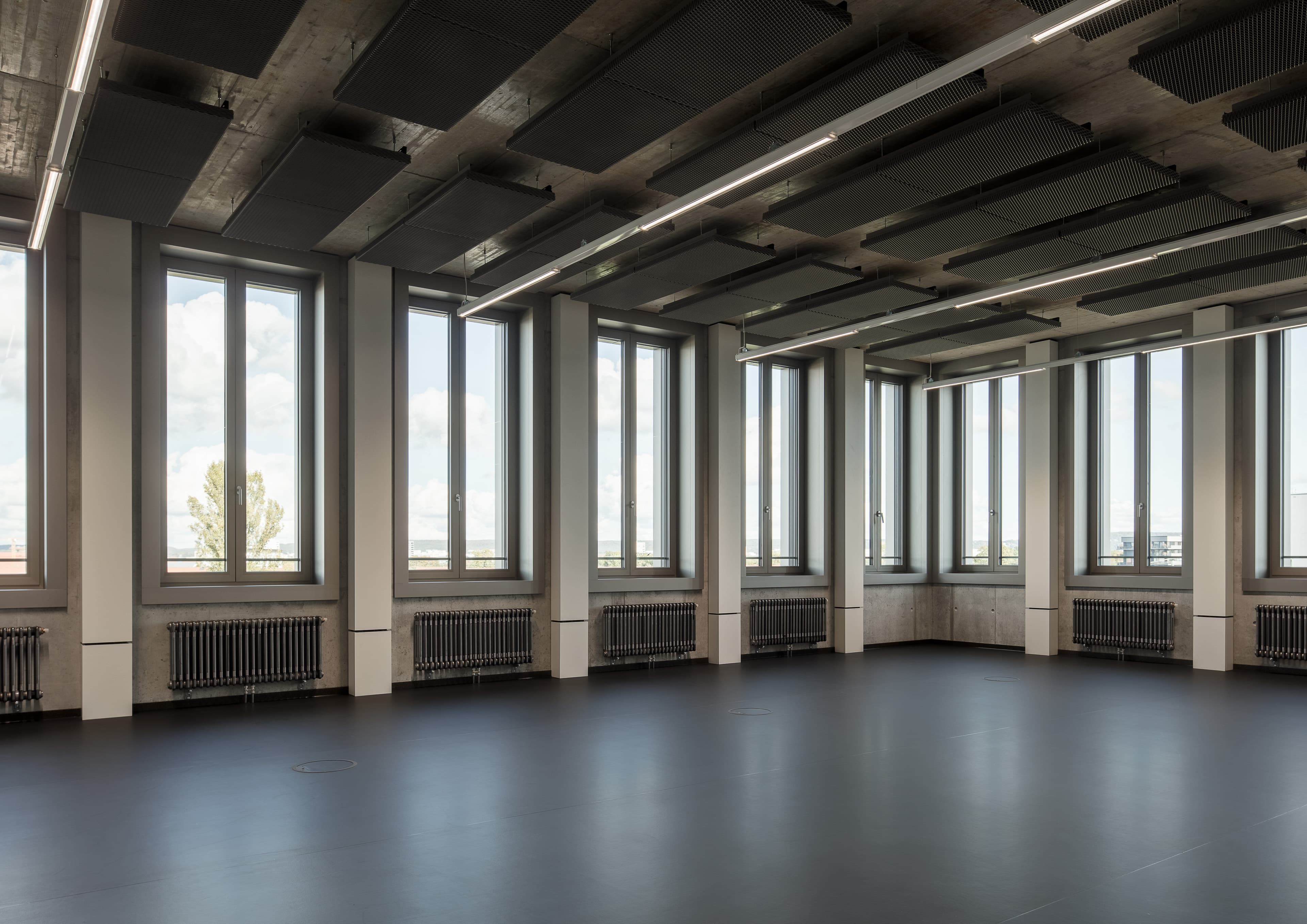 Großzügiger Raum mit hohen Fensterflächen, sichtbarer Deckenkonstruktion und dunklem Boden.