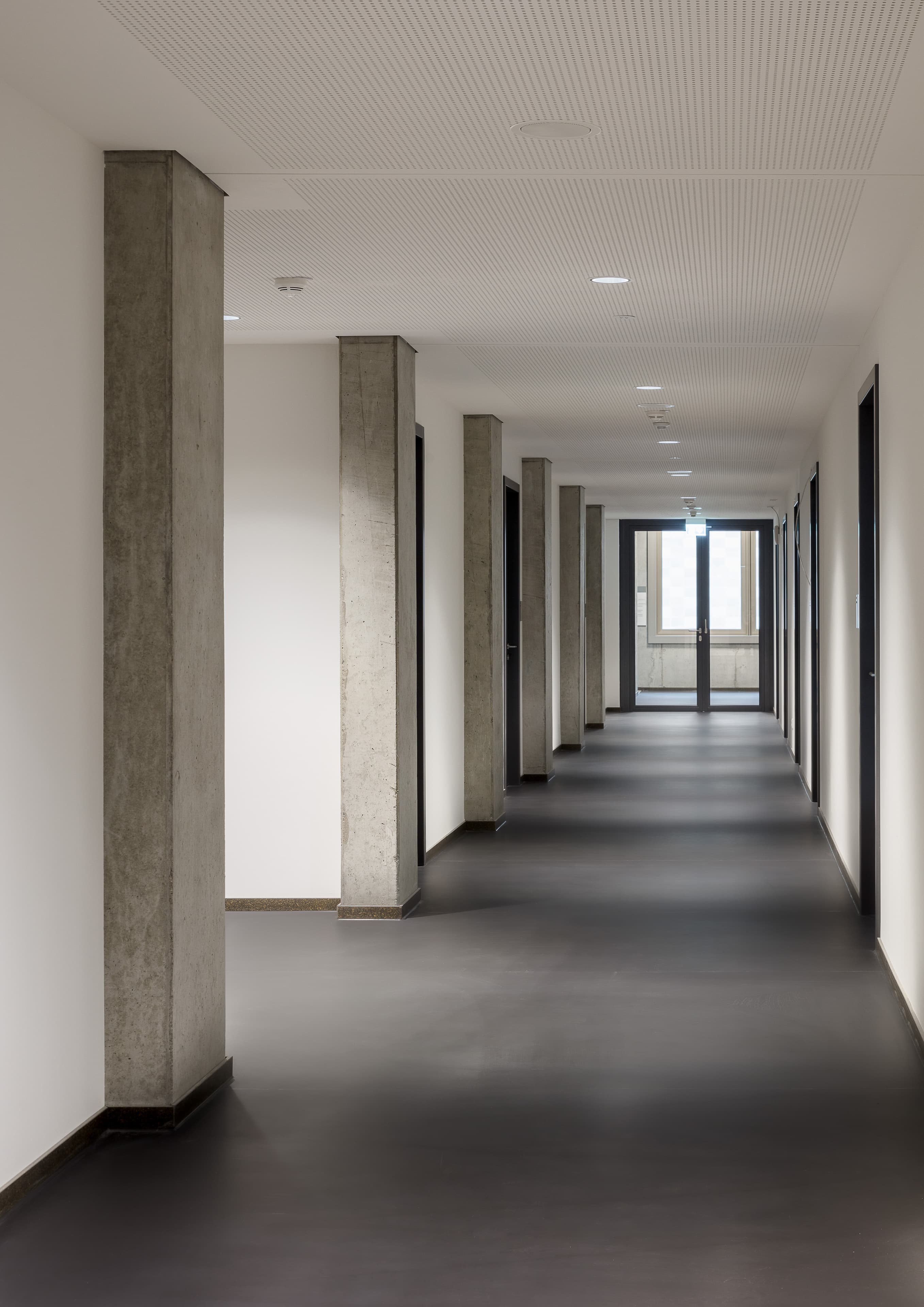 Langer heller Flur mit Sichtbetonstützen, dunklem Boden und Tageslicht am Gangende.