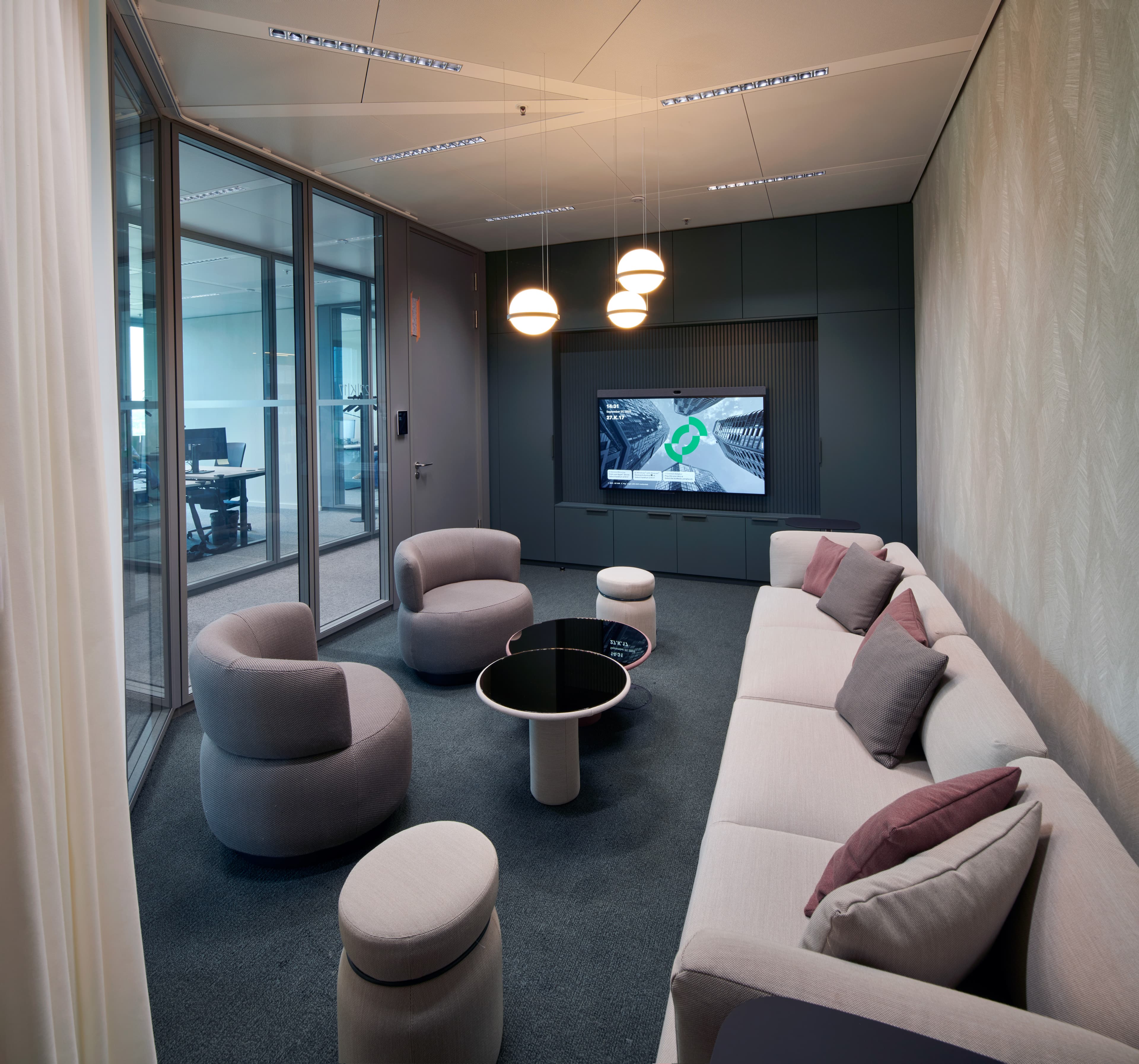 Lounge mit Sofa, Sesseln, runden Tischen, Pendelleuchten und Bildschirm an einer dunklen Wand.
