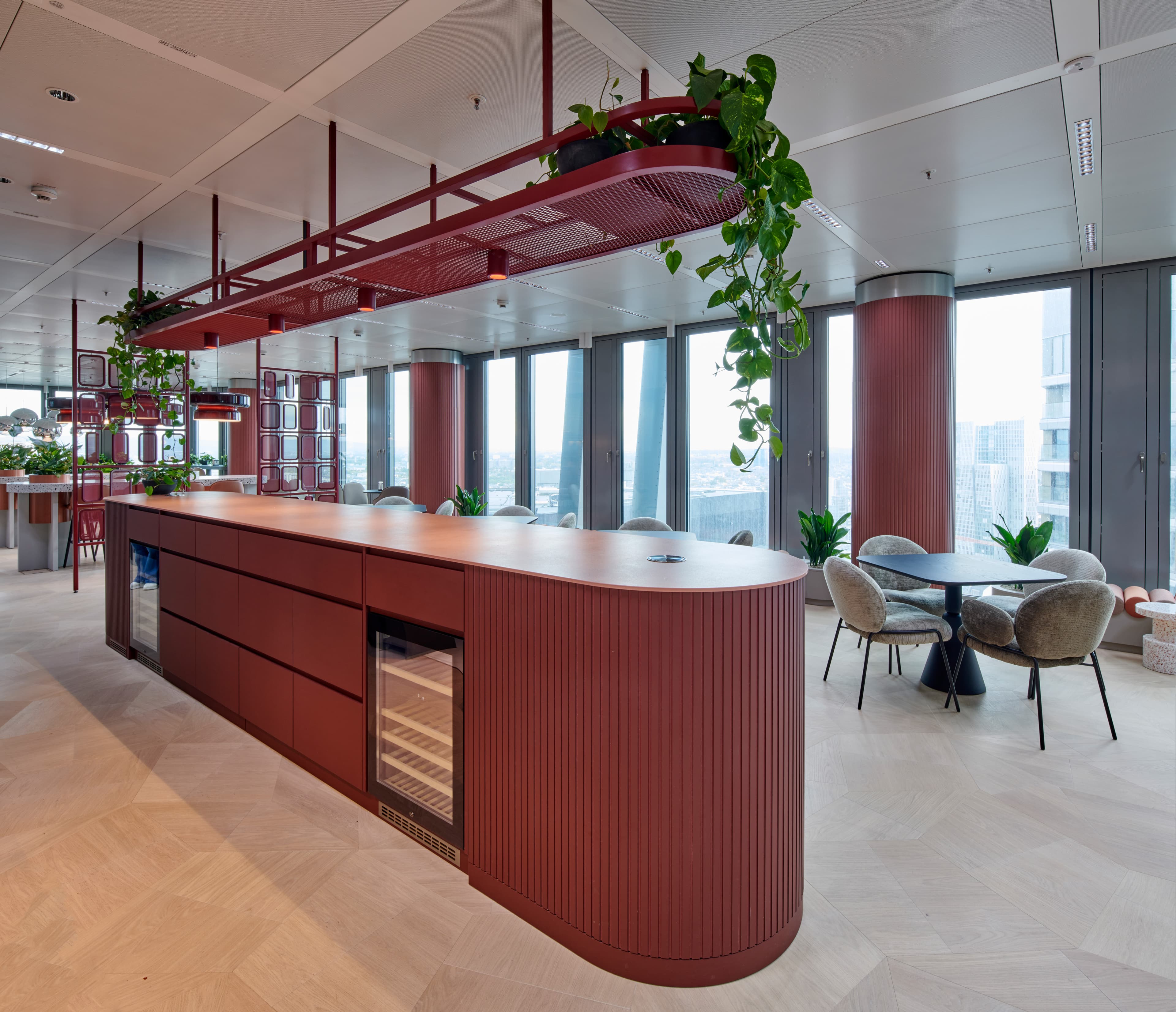 Offene Bürolounge mit dunkelroter Kücheninsel, hängenden Pflanzen, Sitzgruppen und großen Fensterfronten.