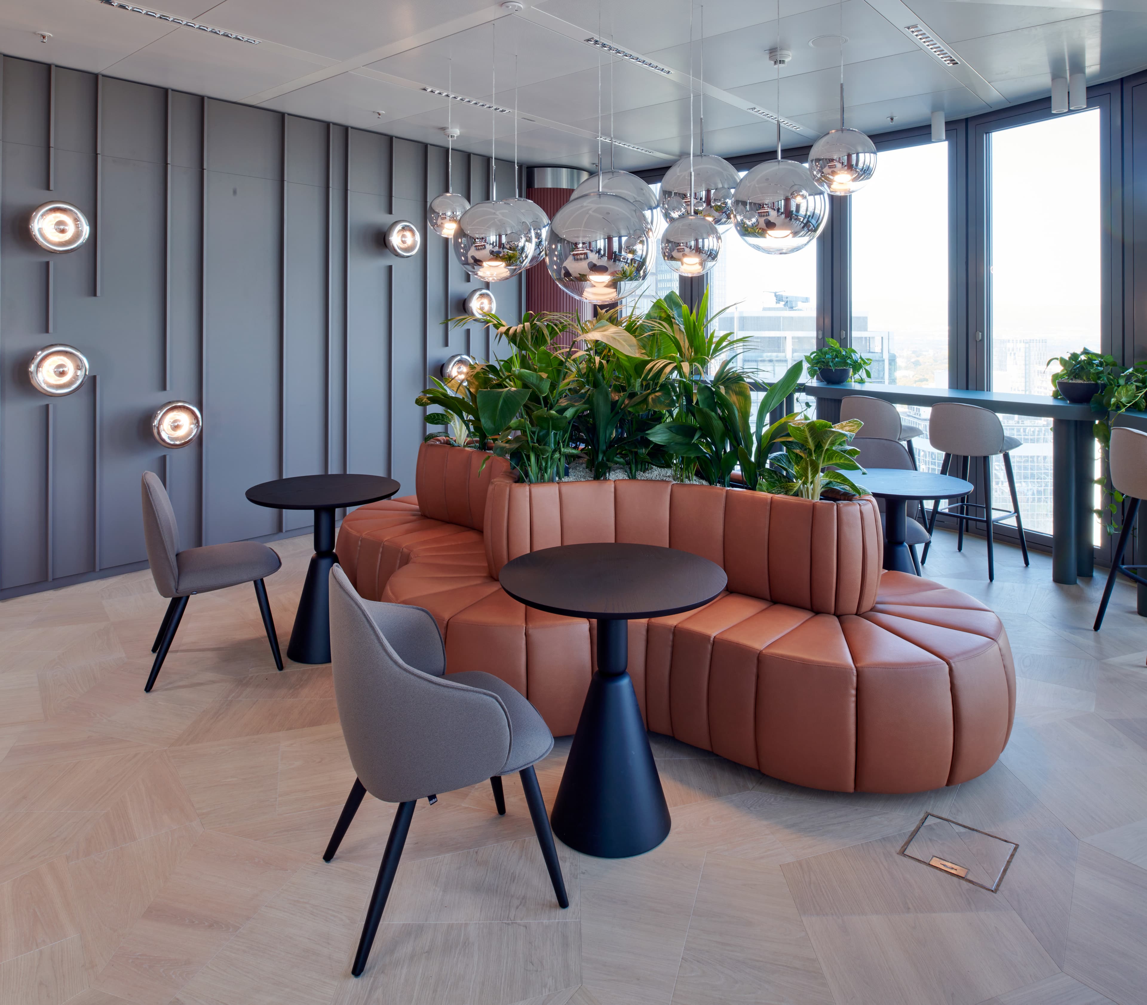 Bürolounge mit rundem, gepolstertem Sofa, integrierten Pflanzen und spiegelnden Pendelleuchten.