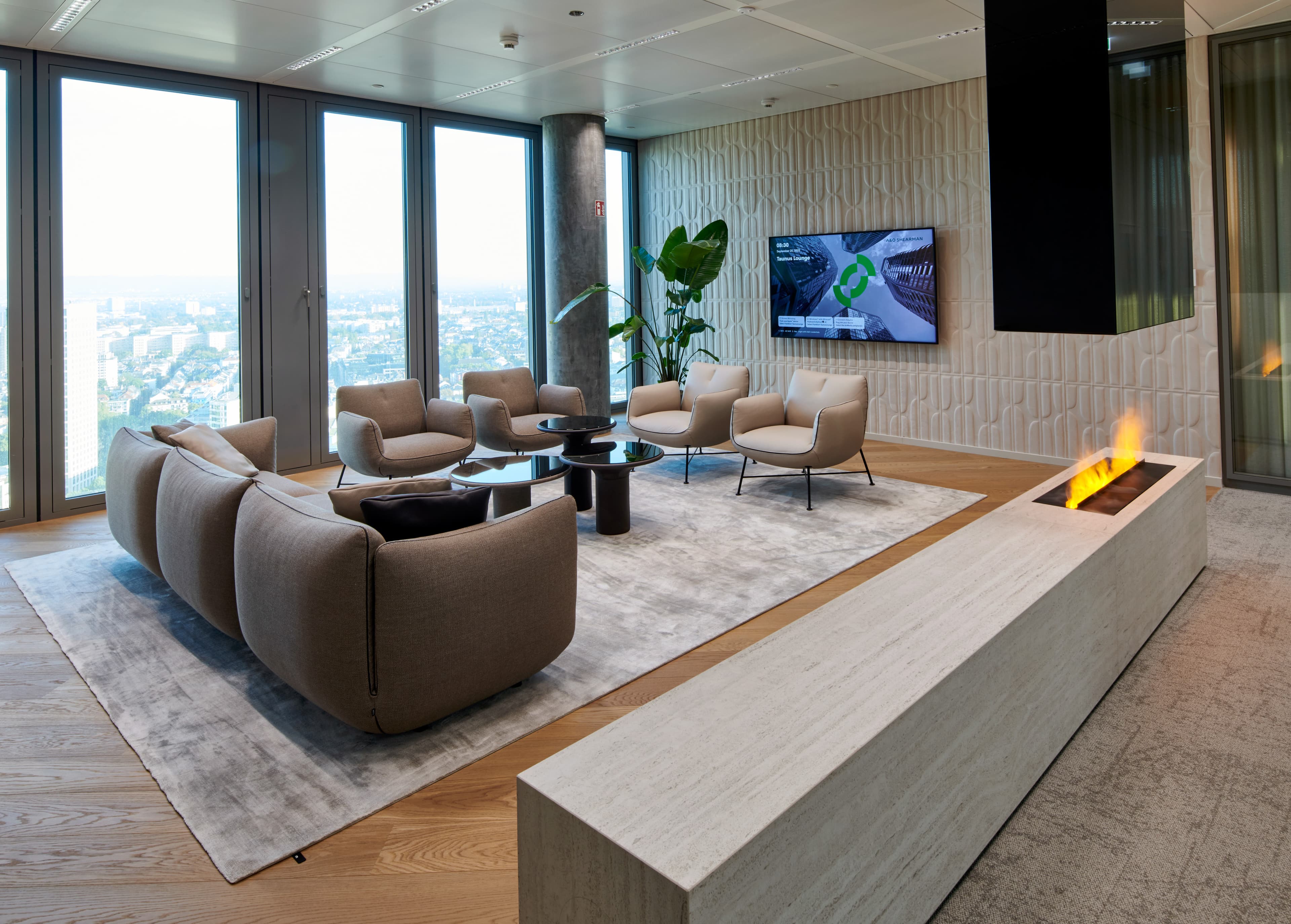 Lounge mit Sesseln, großem Teppich, Bildschirm und offenem Kamin vor Fensterfront mit Stadtblick.