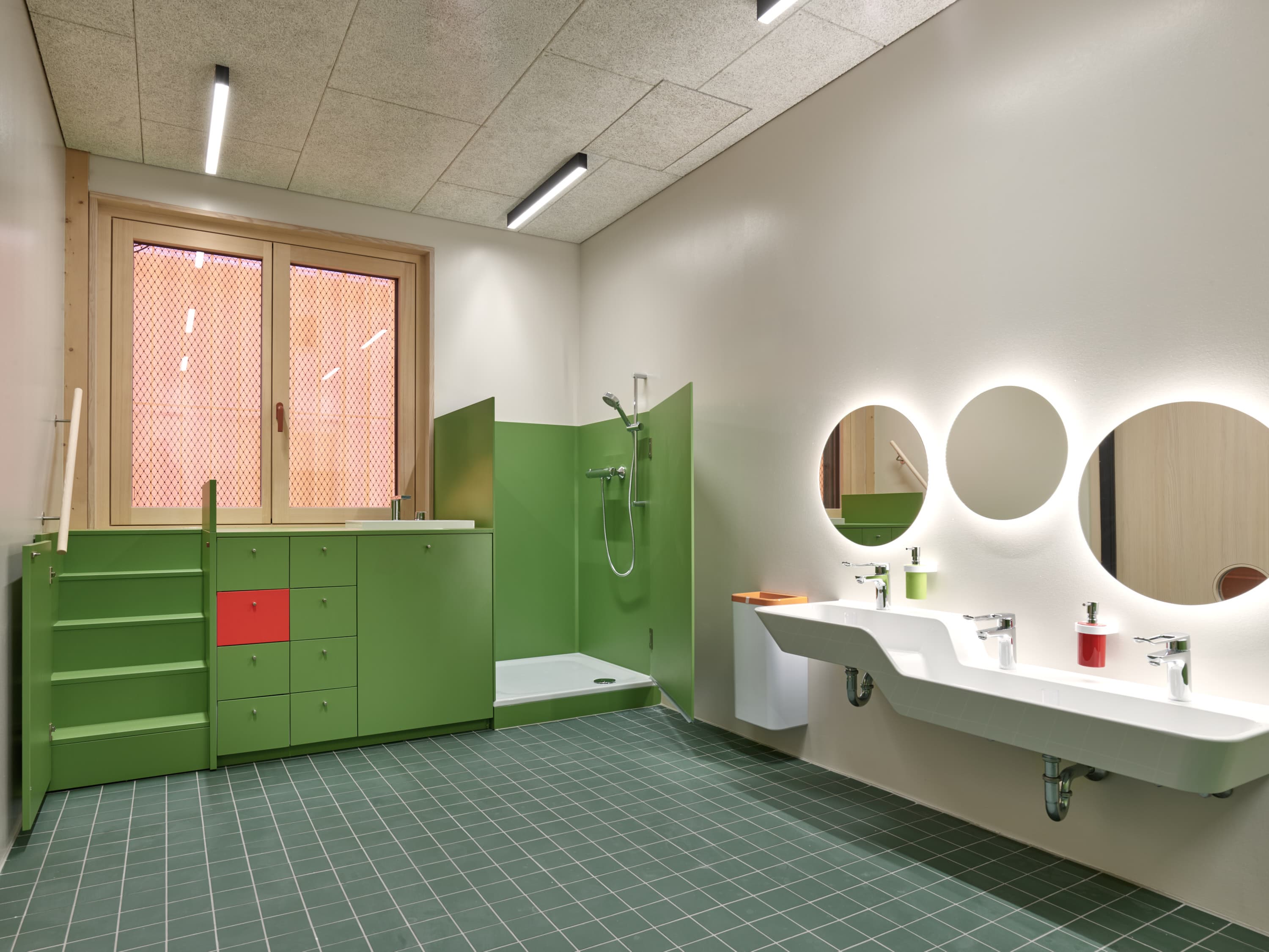 Barrierefreies Kinderbad mit grünen Schränken, Dusche, Waschbecken und runden Spiegeln.