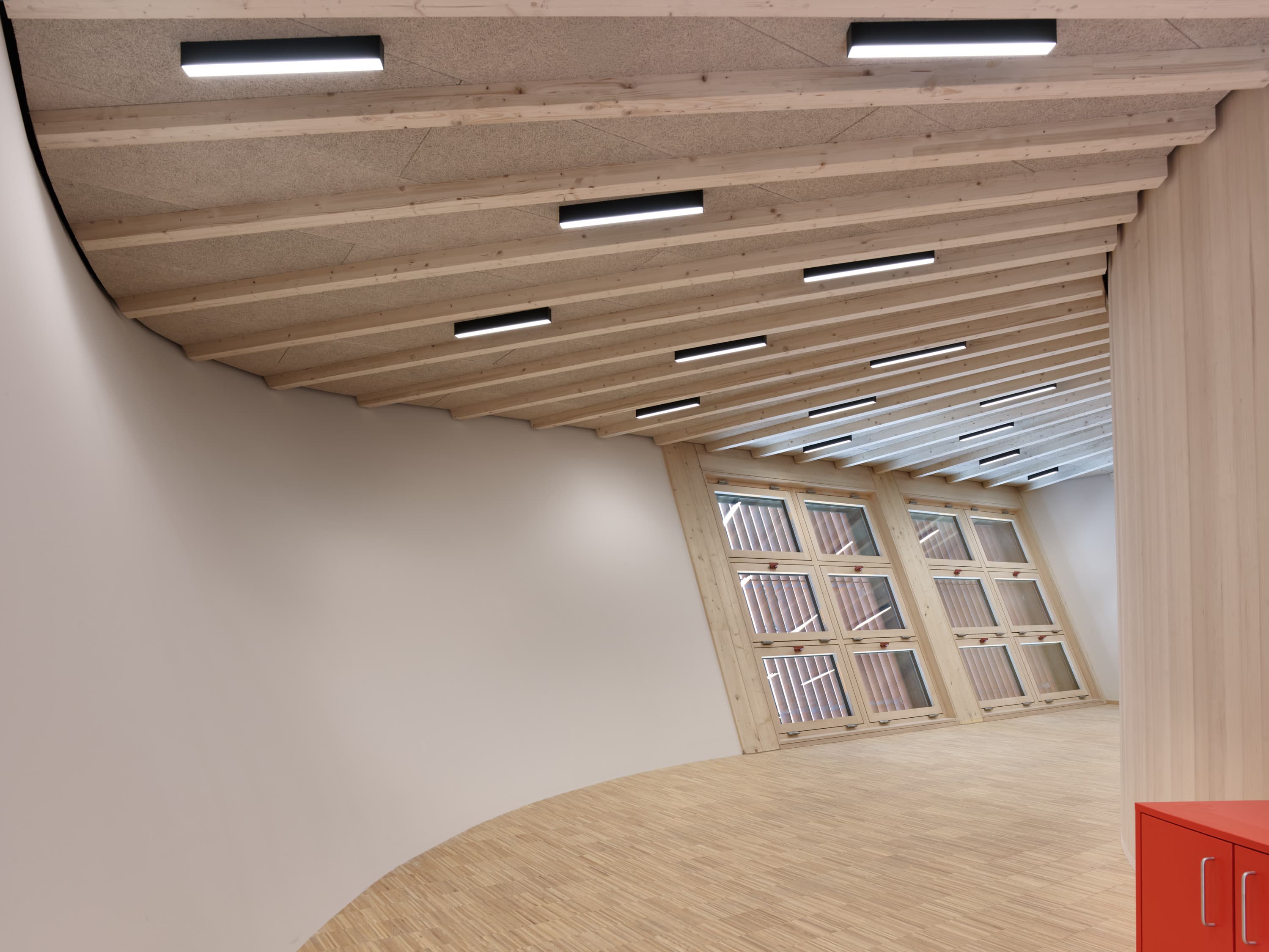 Leerer, geschwungener Innenraum mit Holzbalkendecke, Parkettboden und großen Fensterfronten.