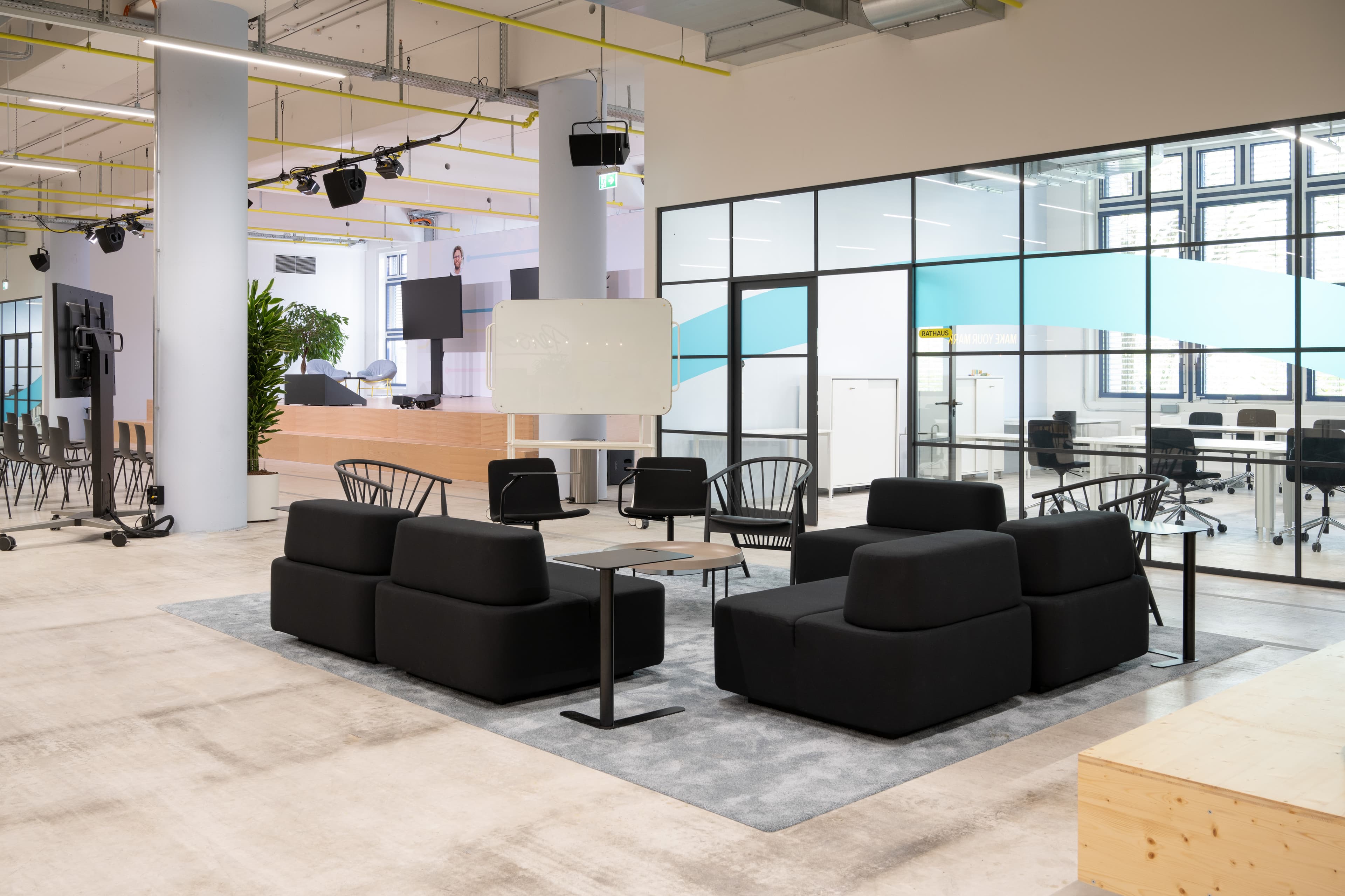 Offene Büro-Lounge mit schwarzen Sesseln, Whiteboard und verglastem Besprechungsraum mit blauer Wellen-Grafik.