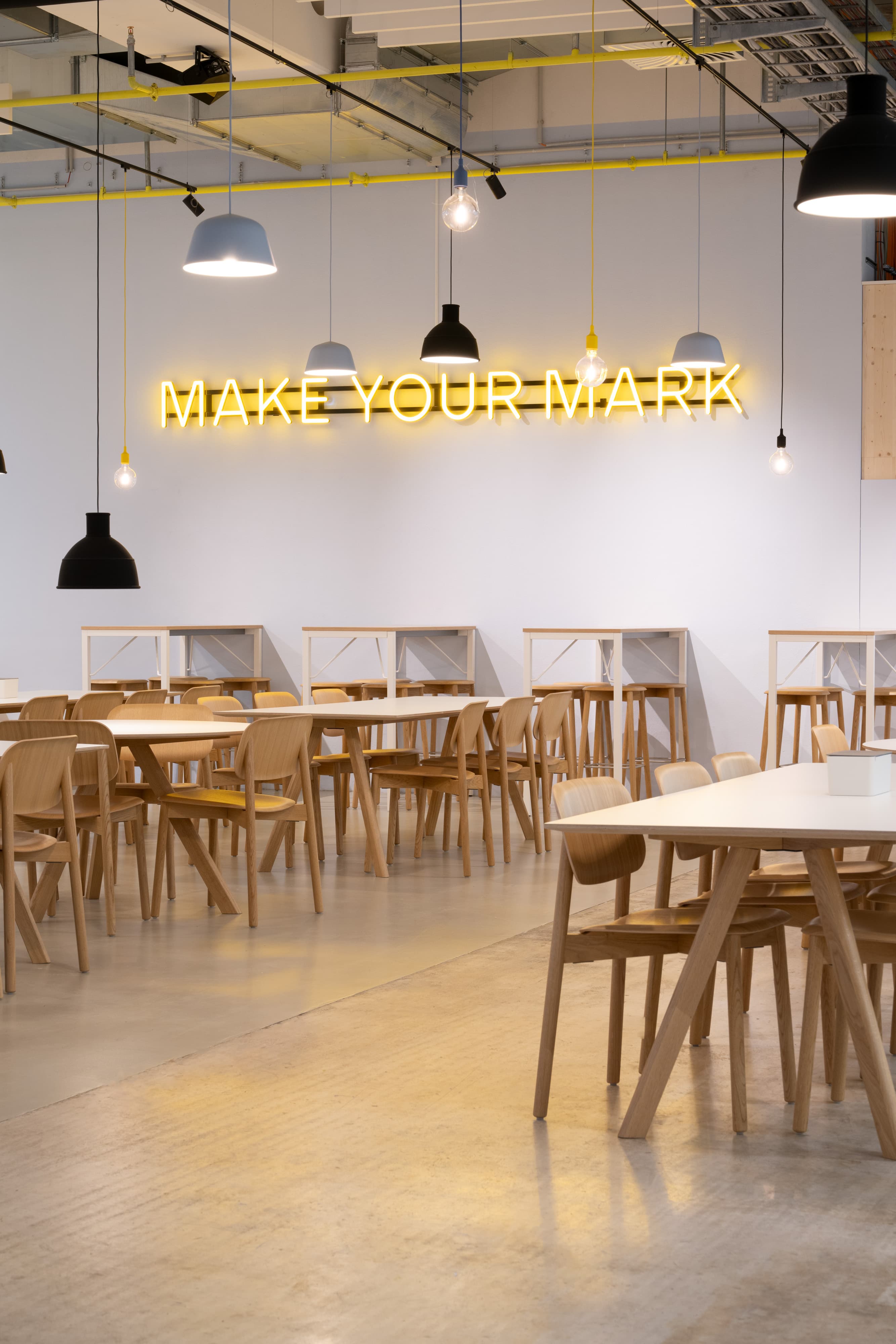 Holztische und Stühle unter Pendelleuchten vor gelbem „MAKE YOUR MARK“-Schriftzug.