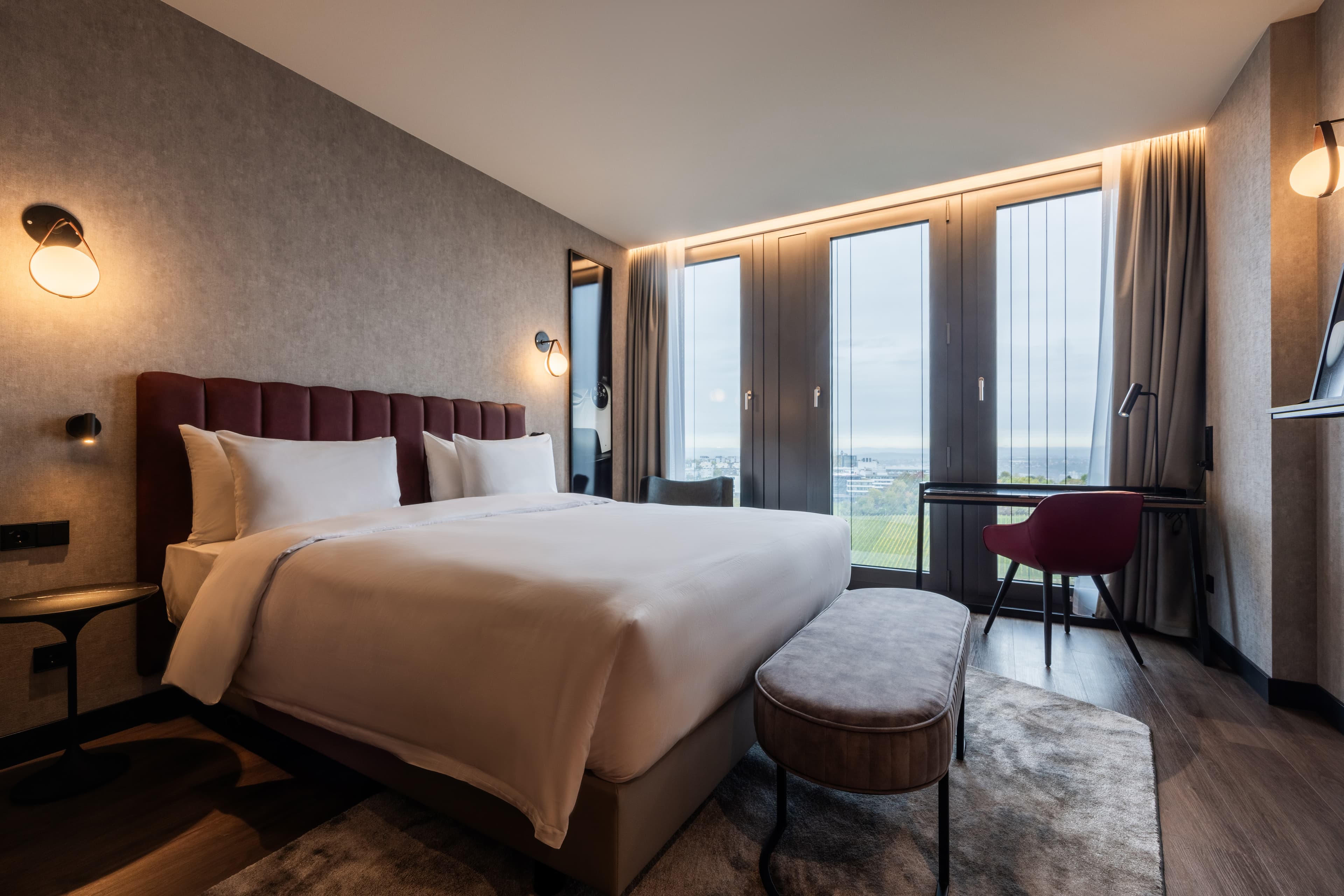 Modernes Hotelzimmer mit Doppelbett, Sitzbank, Schreibtisch und bodentiefen Fenstern.