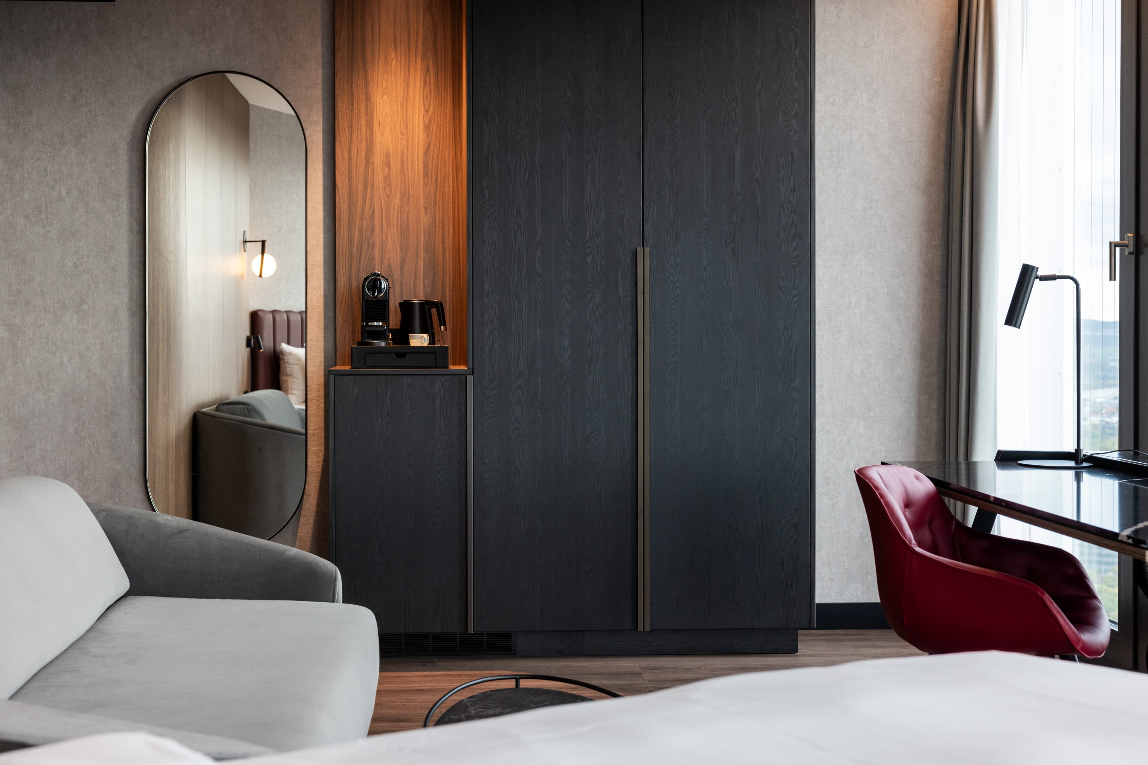 Hotelzimmer mit grauem Sofa, dunklem Kleiderschrank, großem Spiegel und Schreibtisch mit rotem Sessel.