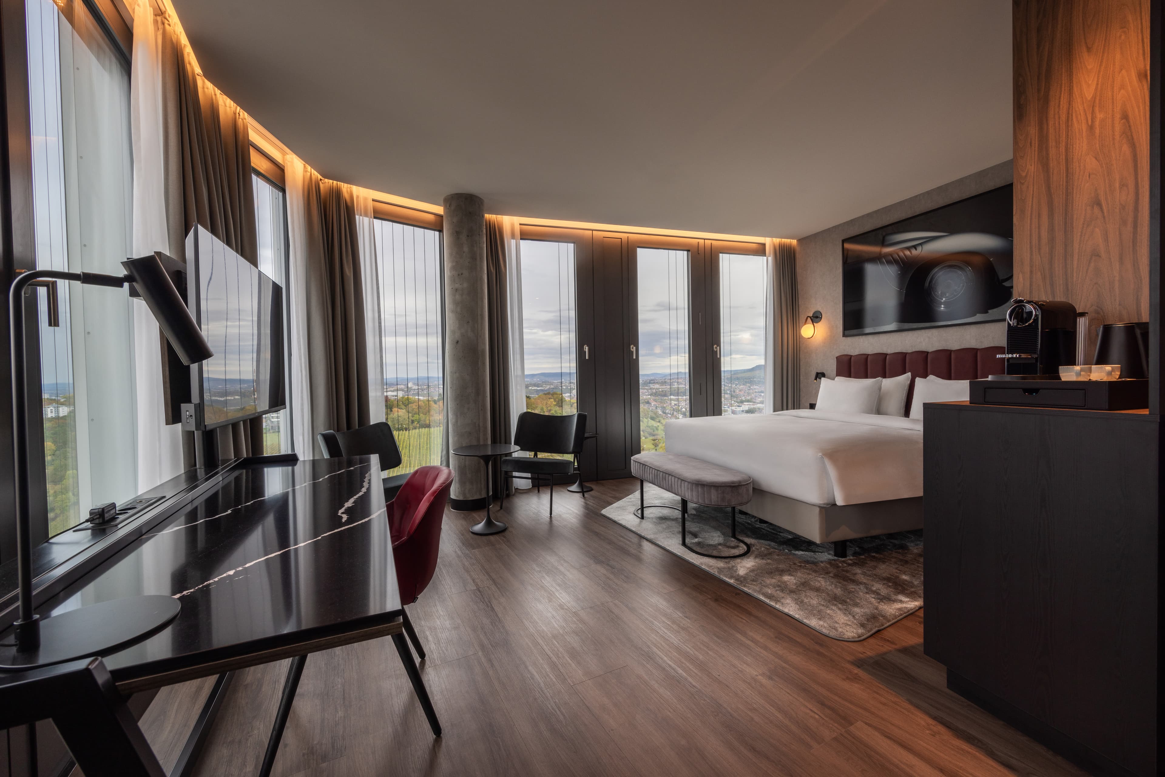 Modernes Hotelzimmer mit Doppelbett, Schreibtisch, Sitzecke und bodentiefen Fenstern mit Stadtblick.
