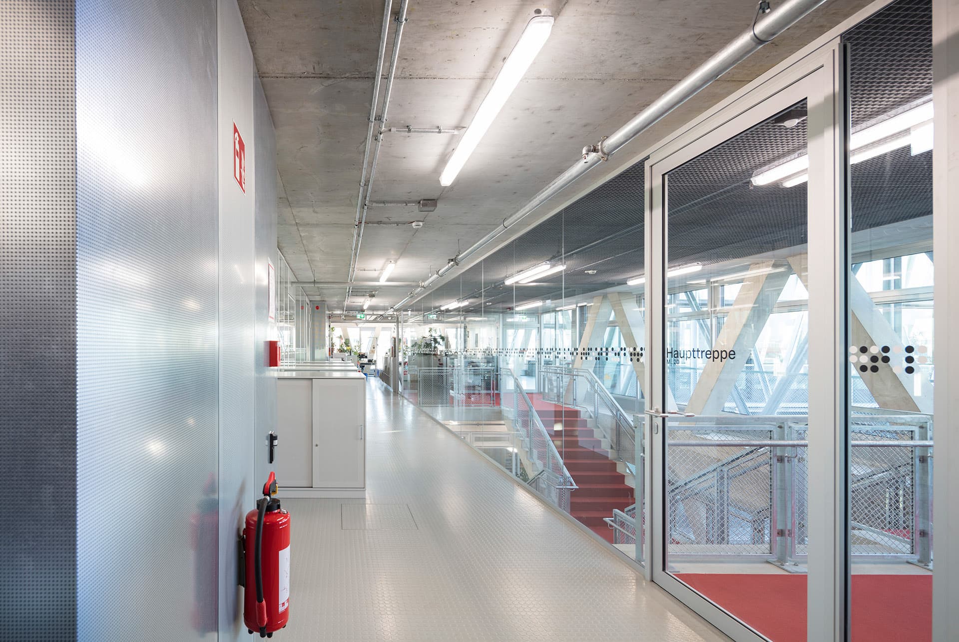Heller Flur mit Glaswänden, sichtbaren Installationen und Blick auf eine rote Treppe.