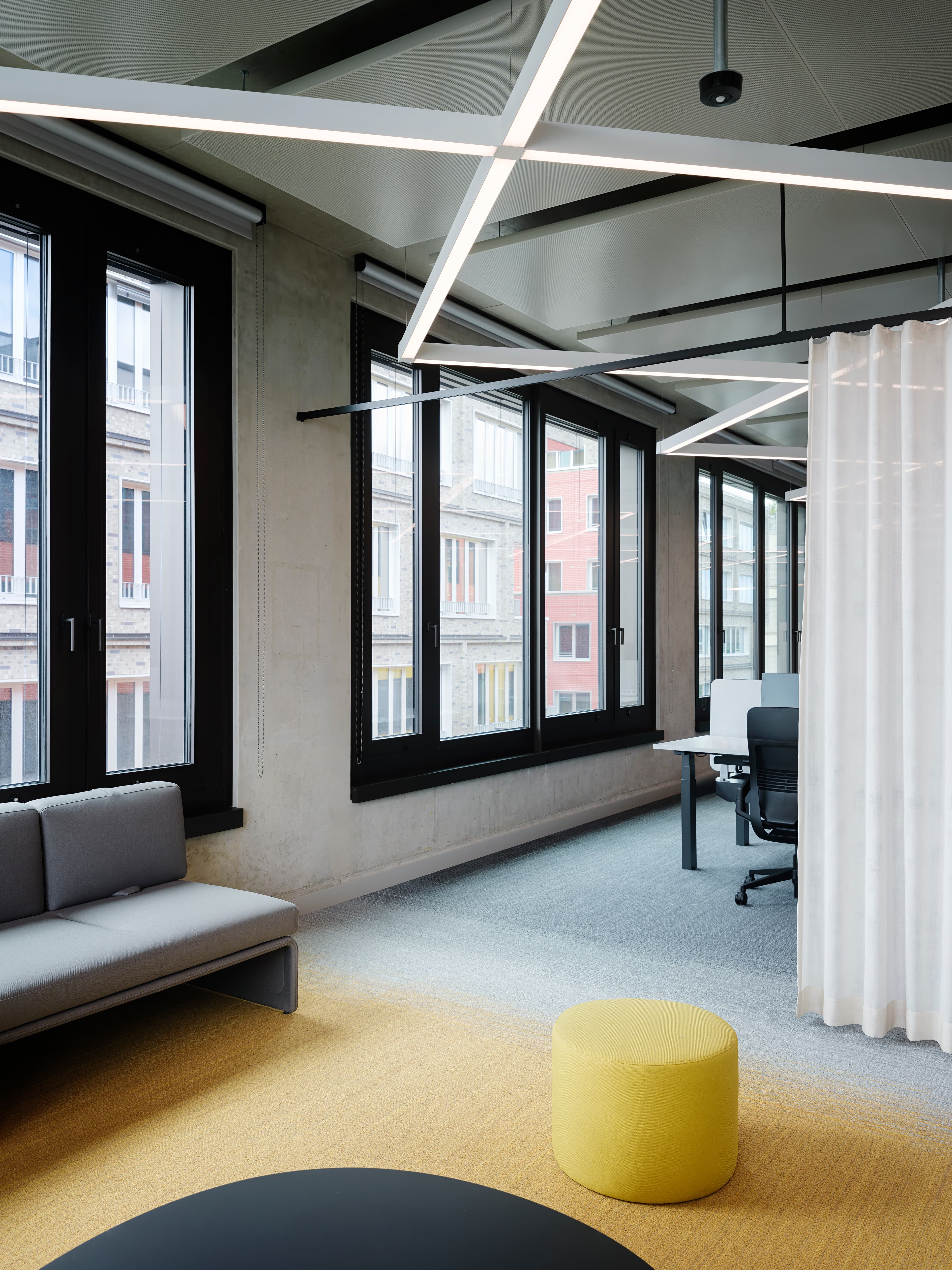 Heller Bürobereich mit Sofa, Schreibtisch, gelbem Hocker und bodentiefen Fenstern.