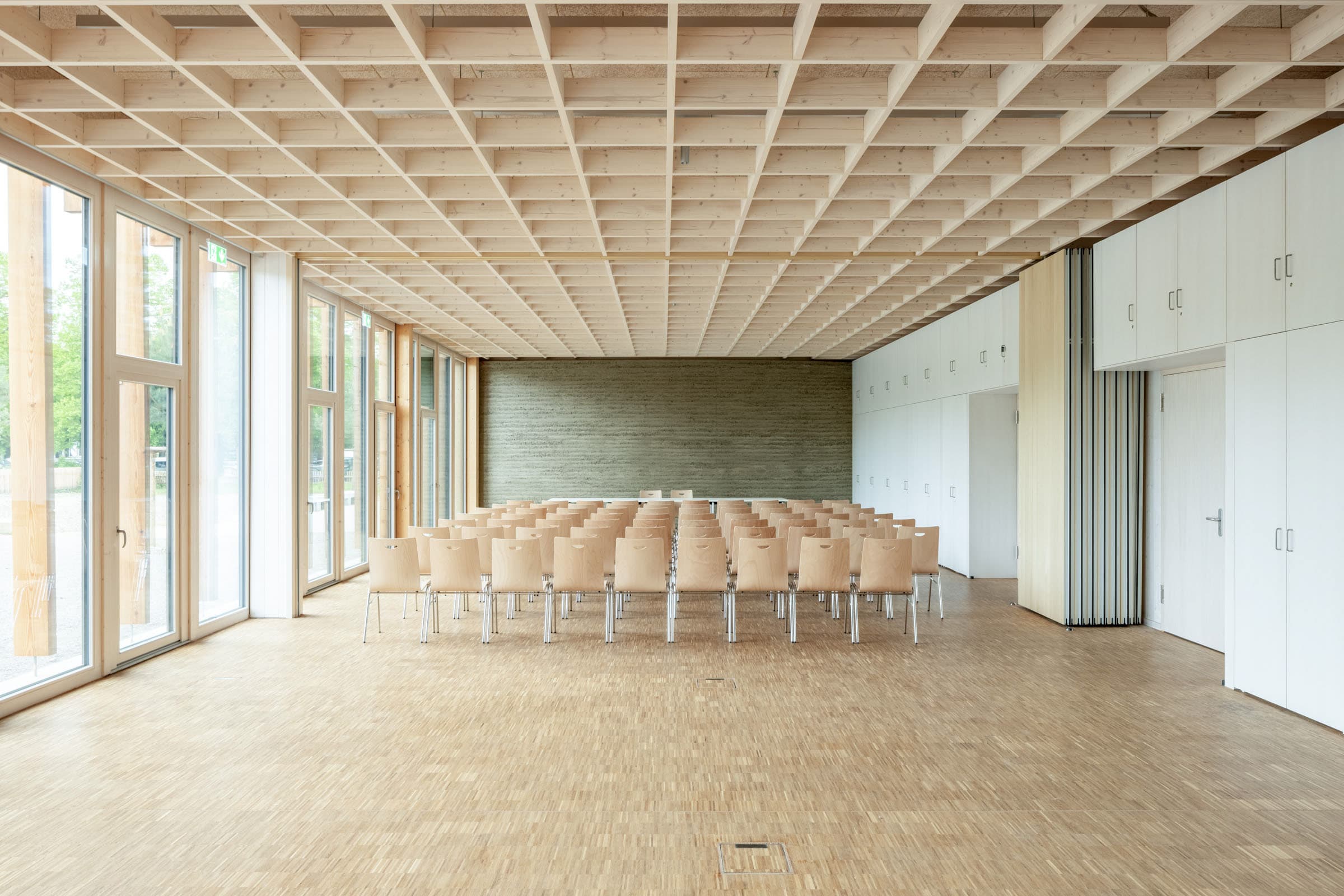 Heller Veranstaltungsraum mit Holzrasterdecke, langen Fenstern und Holztischen.