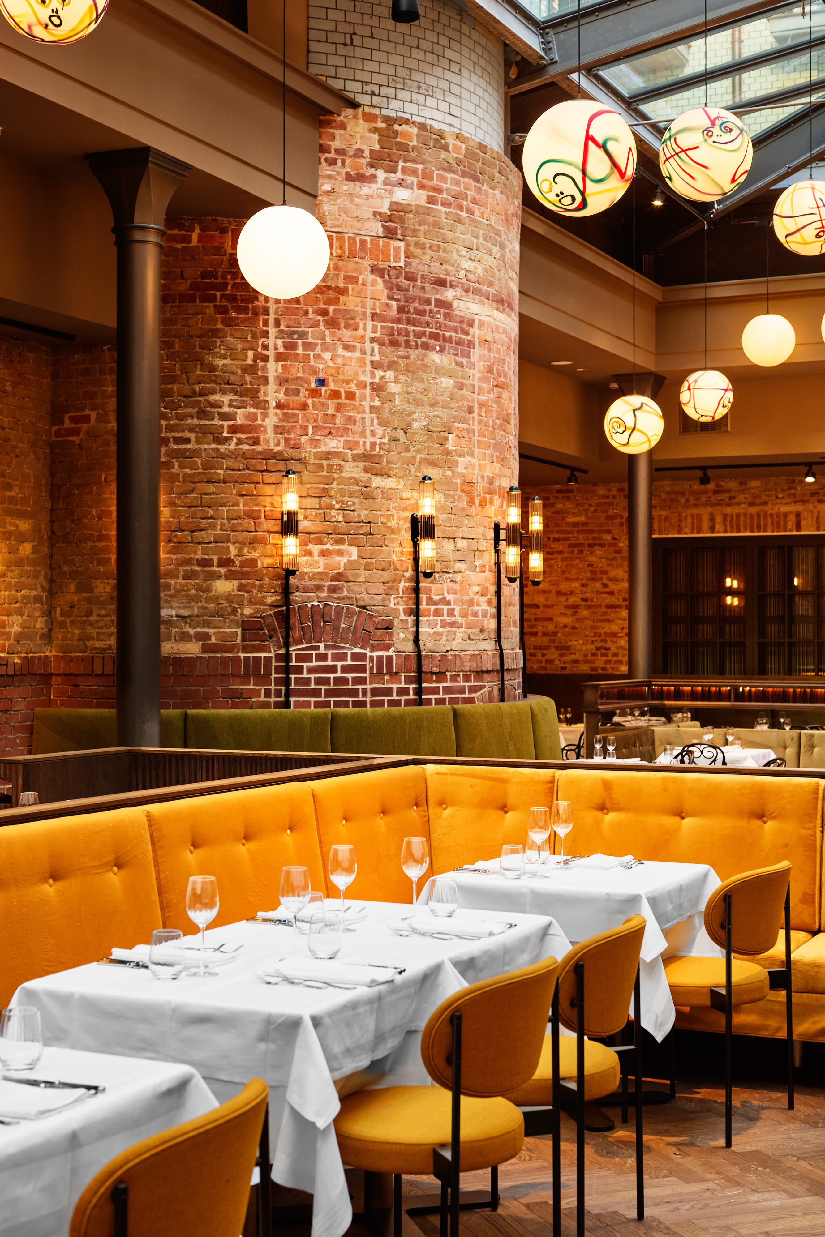 Restaurant mit gelben Sitzbänken, gedeckten Tischen und runden Pendelleuchten vor Ziegelwänden.