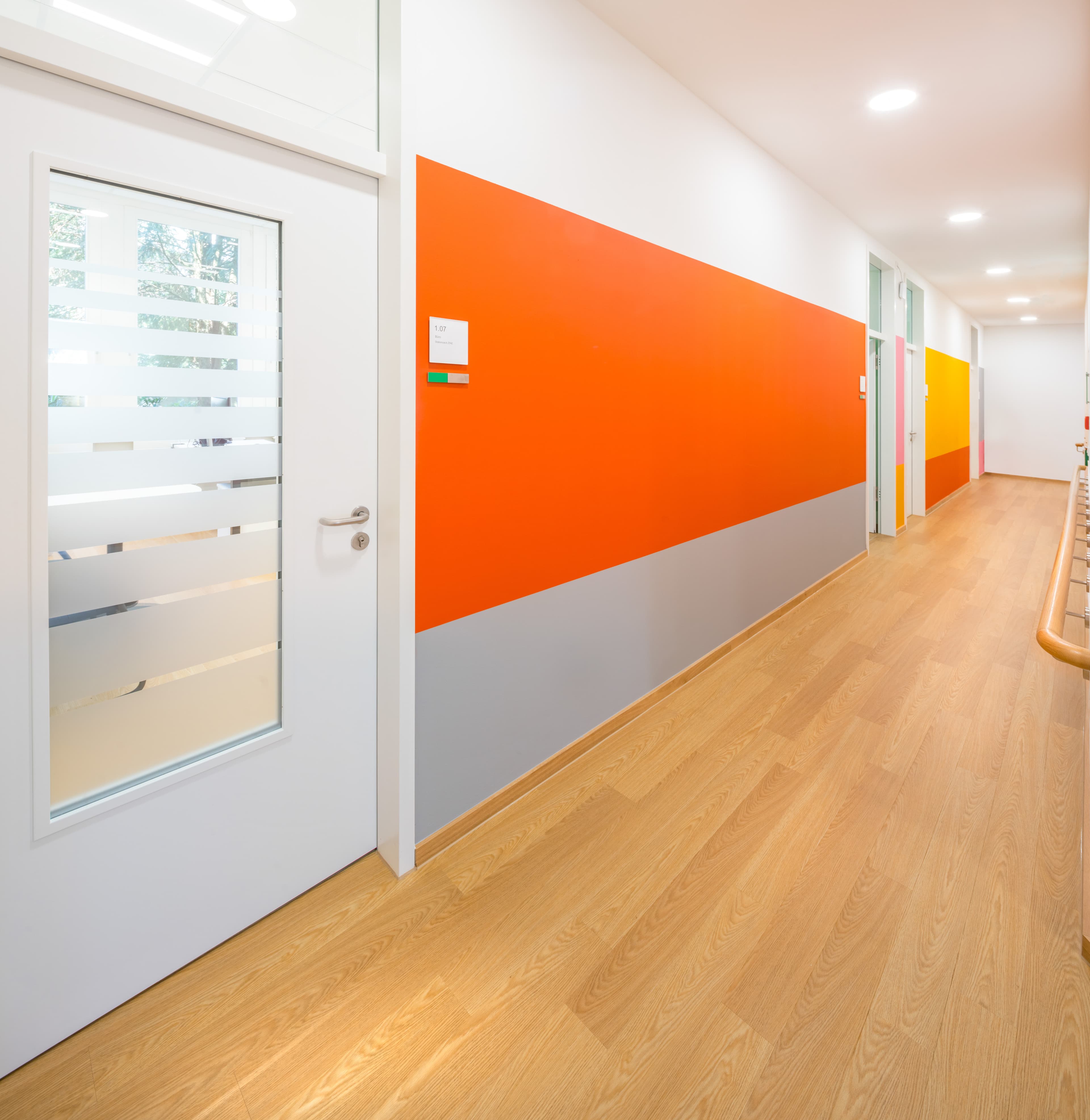 Heller Flur mit Holzboden, Glastür und orange-grauer Wandgestaltung.
