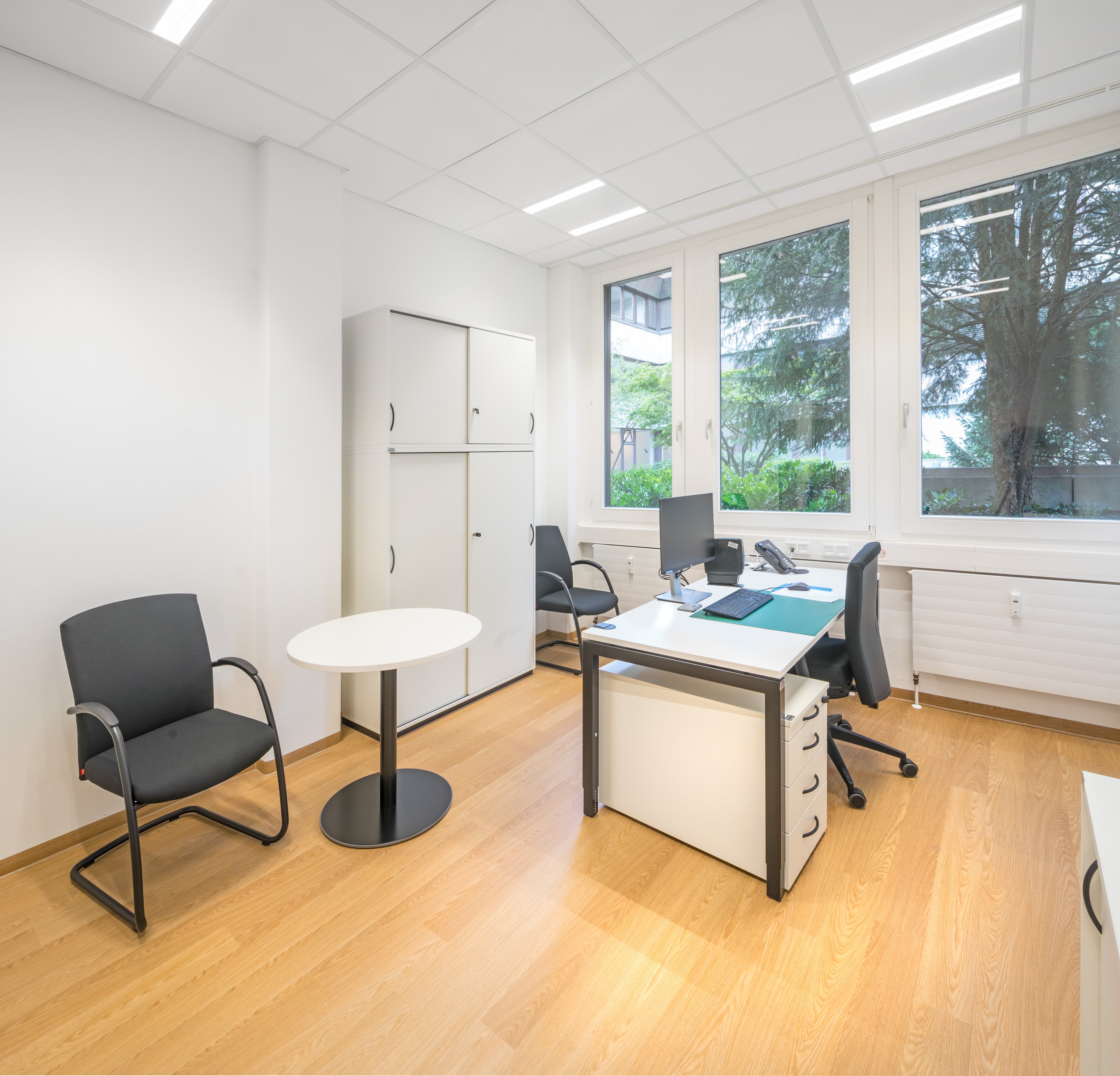 Helles Büro mit Schreibtisch, zwei Besucherstühlen, Schrank und großen Fenstern.