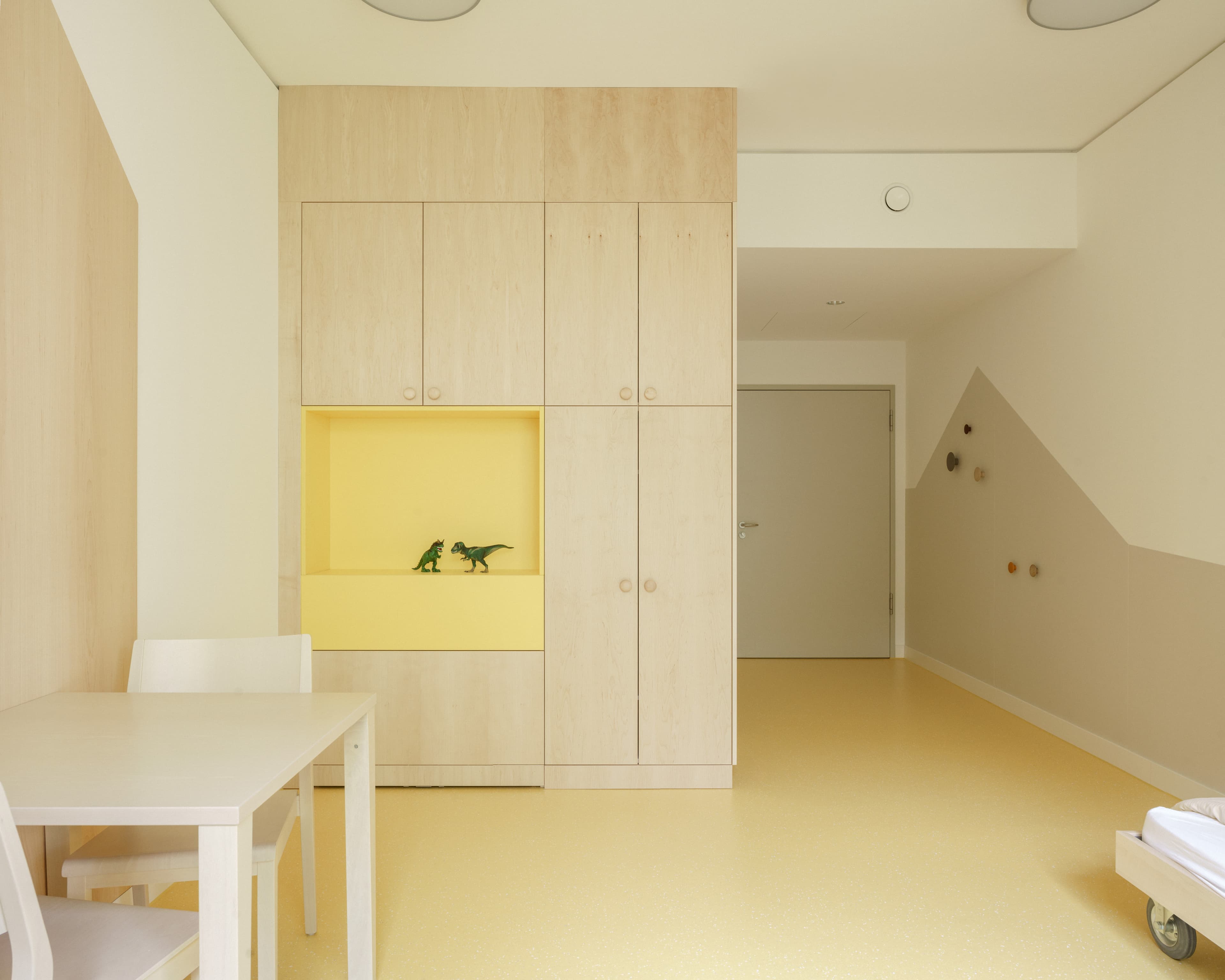 Helles Zimmer mit großem Holzschrank, gelbem Regal, Tisch und einer Kletterwand an der Zimmerwand.