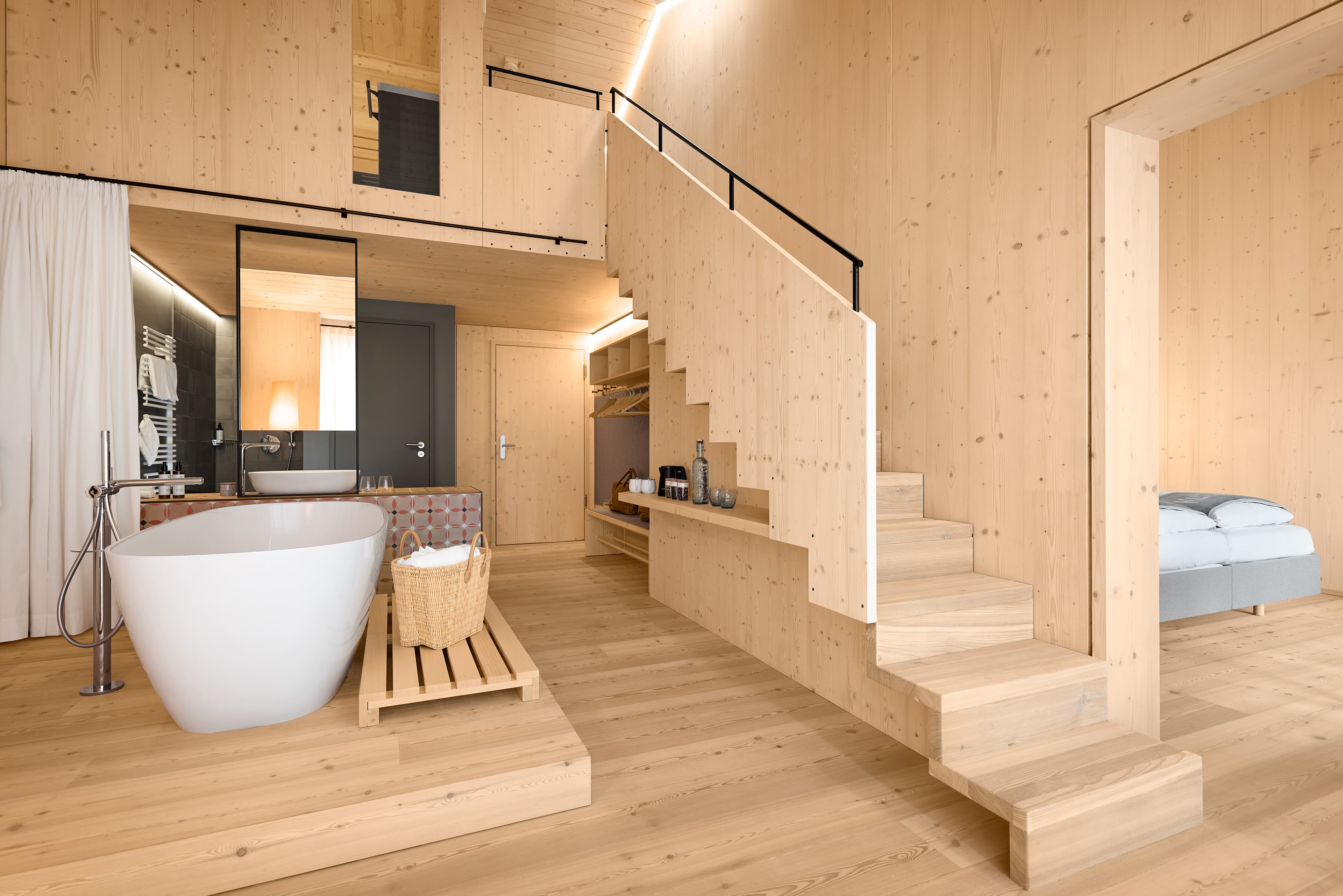 Helles Hotelzimmer mit viel naturbelassenem Holz, freistehender Badewanne, offener Treppe und Blick in den angrenzenden Schlafbereich.