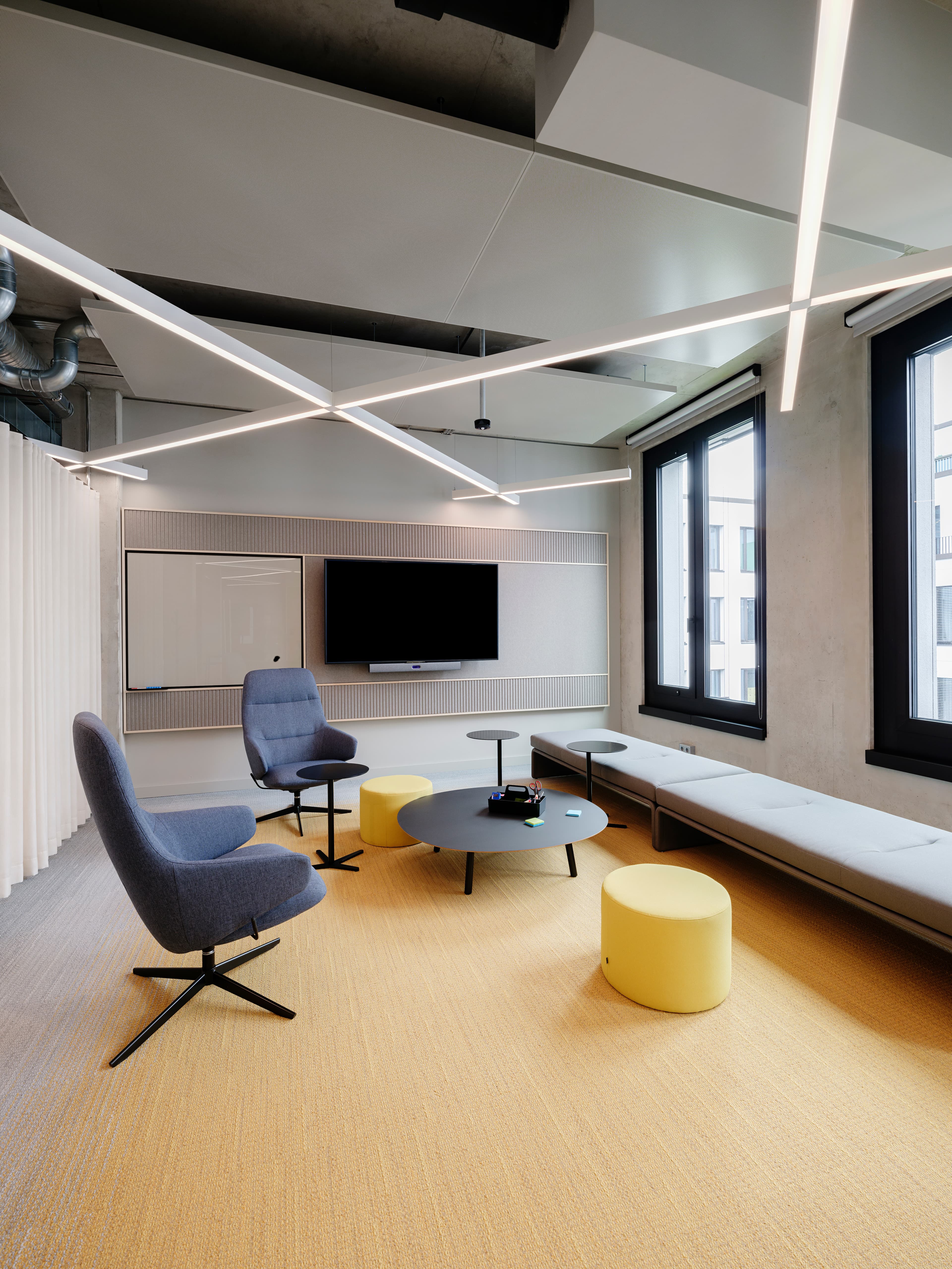 Büro-Loungebereich mit Sesseln, kleinem Tisch und gelbem Hocker vor großen Fenstern.