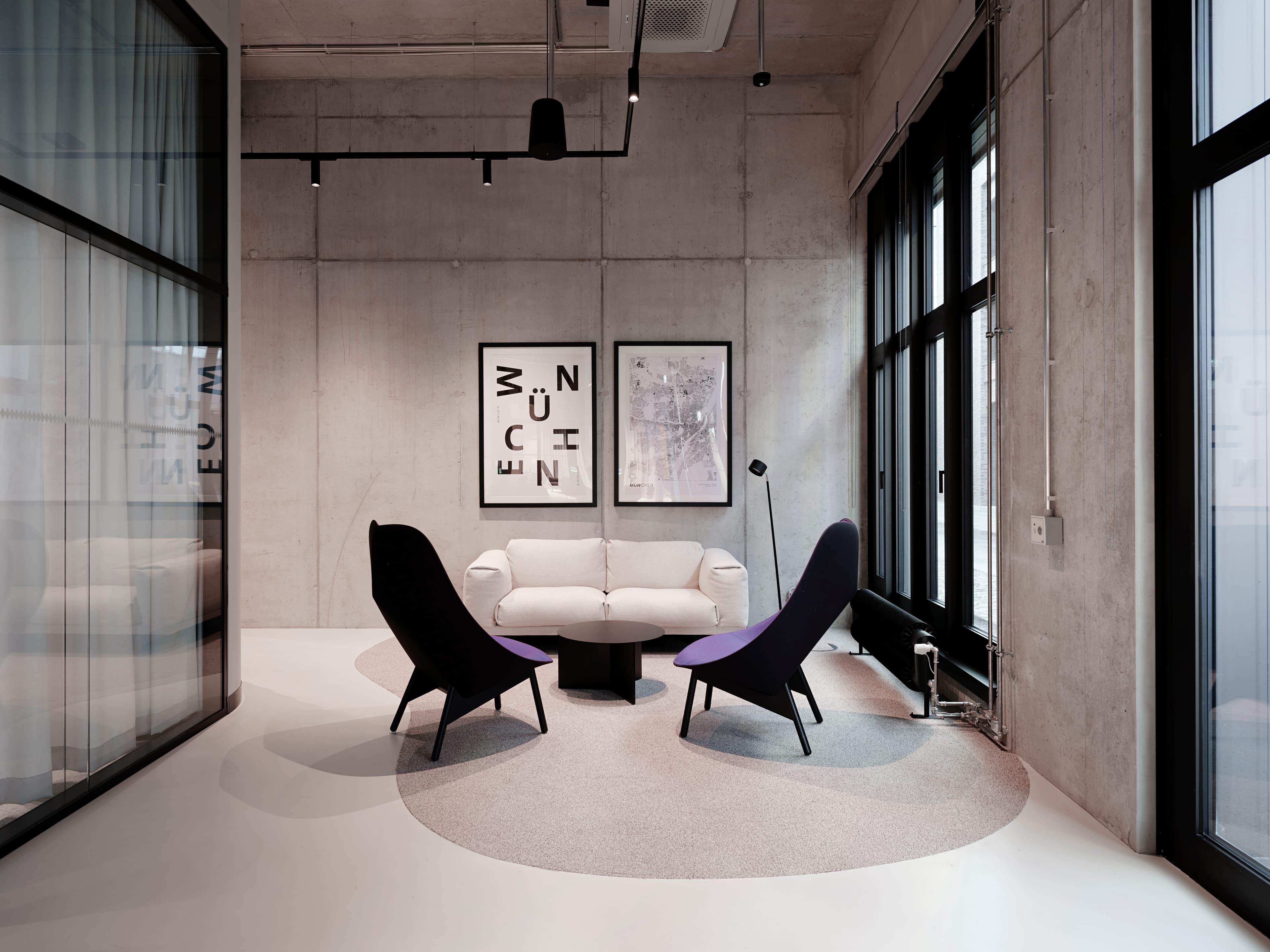 Lounge mit zwei schwarzen Sesseln, Sofa und runder Teppichfläche vor Betonwänden.