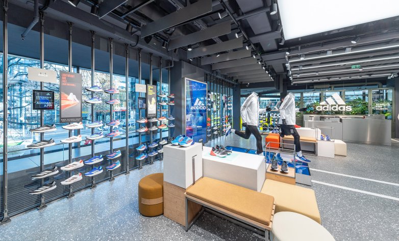 Sitzgelegenheiten in Ausstellungsfläche, Service-Theke mit Adidas Logo im Hintergrund, daneben große Fensterfront mit Laufschuhen in offenen Regalen