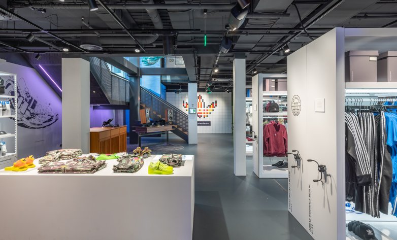 Hell gestalteter Einkaufsbereich mit Regalen, daneben Servicetheke und DJ-Pult in Holzoptik, im Hintergrund buntes Adidaslogo
