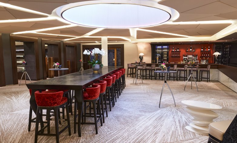Stilvolle Bar- und Loungefläche mit zentralem Hochtisch und eleganten, roten Barhockern. Die moderne Deckenbeleuchtung mit kreisförmigem Lichtelement sorgt für angenehme Atmosphäre. Im Hintergrund eine gut sortierte Bar mit Weinregalen, Sitznischen und dekorativen Blumenarrangements – ideal für Empfänge, Networking oder entspannte Abende.