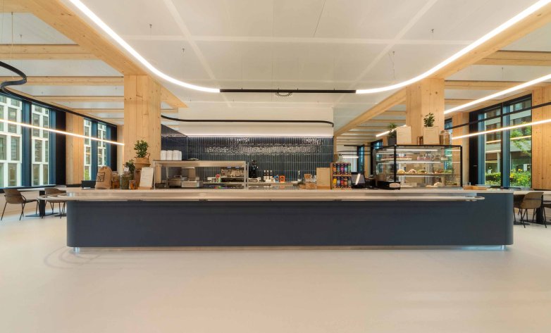Modern gestaltete Cafeteria mit klaren Linien, viel Tageslicht und hochwertigen Materialien: Der zentrale Tresen lädt zum Verweilen ein und verbindet Funktionalität mit einem einladenden Ambiente für Mitarbeitende und Gäste.
