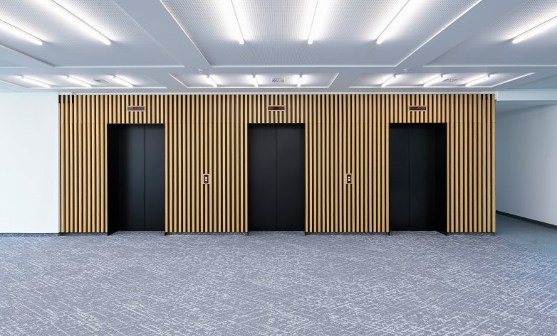Moderner Aufzugsbereich mit drei schwarzen Türen, eingefasst in eine elegante Wandverkleidung aus vertikalen Holzlamellen. Der helle Teppichboden und die gleichmäßige Deckenbeleuchtung schaffen eine freundliche und klare Atmosphäre.