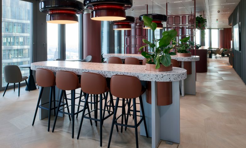 Modern eingerichteter Aufenthaltsbereich mit hohen Tischen aus hellem Terrazzo und bequemen Barhockern in warmem Braunton. Große Fenster lassen viel Tageslicht herein, während grüne Pflanzen und stilvolle Deckenleuchten eine angenehme Atmosphäre schaffen. Das elegante Design kombiniert natürliche Materialien mit modernen Akzenten – ideal für Pausen, Meetings oder entspannte Gespräche.