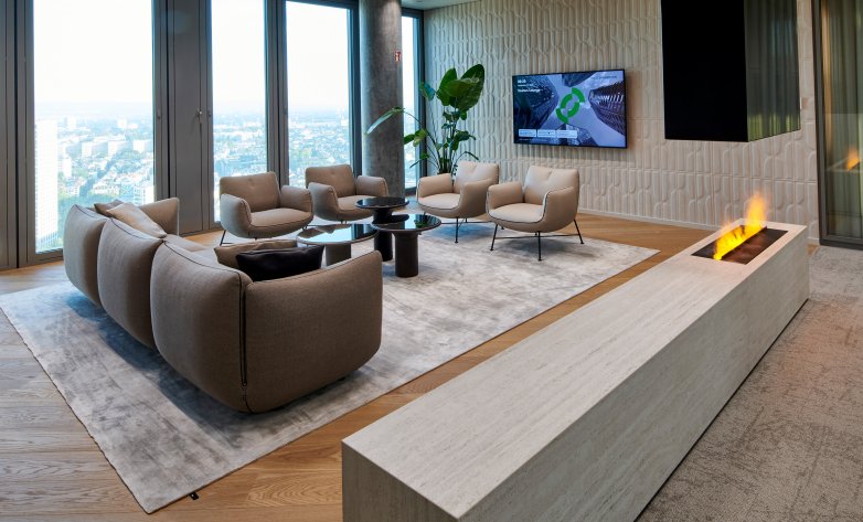 Eleganter Loungebereich mit modernen Sofas und Sesseln in neutralen Farbtönen, arrangiert um einen runden Couchtisch auf einem hellen Teppich. Ein eingebauter Kamin sorgt für warme Atmosphäre, während große Fenster einen beeindruckenden Blick über die Stadt bieten. Dezente Wandstrukturen, Pflanzen und ein Bildschirm verleihen dem Raum eine stilvolle und zugleich gemütliche Note – ideal für entspannte Meetings oder Gespräche.