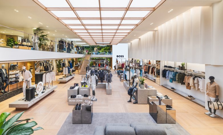 Geräumiger, modern gestalteter BOSS Store mit hellen Holzböden und einer beleuchteten Decke in Rasteroptik. In der Mitte stehen stilvolle Mannequins mit Sport- und Freizeitmode, umgeben von Präsentationstischen und Regalen mit Kleidung und Accessoires. Eine offene Treppe führt zur oberen Etage, wo weitere Kollektionen ausgestellt sind. Die Atmosphäre wirkt hochwertig, einladend und urban.