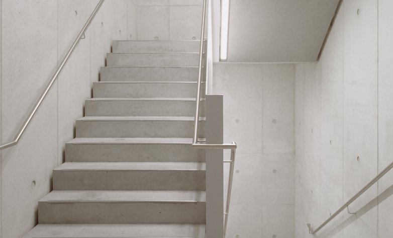Eine schlichte Betontreppe mit Edelstahlgeländern in einem modernen Gebäude. Die klaren Linien und das helle Licht schaffen eine minimalistische, funktionale Atmosphäre. Wände, Stufen und Decke bestehen aus glattem Sichtbeton, was dem Raum ein puristisches, industrielles Erscheinungsbild verleiht.