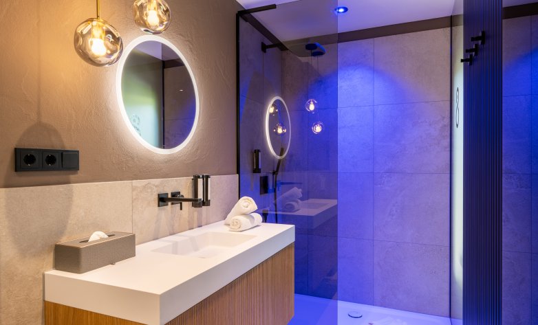 Ein modernes Badezimmer mit elegantem Design: Eine beleuchtete Rundspiegel-Wand, stilvolle Hängelampen und eine begehbare Dusche mit blauer Akzentbeleuchtung schaffen ein luxuriöses Ambiente. Helle Fliesen, dunkle Armaturen und Holzelemente verleihen dem Raum Wärme und zeitlose Eleganz.