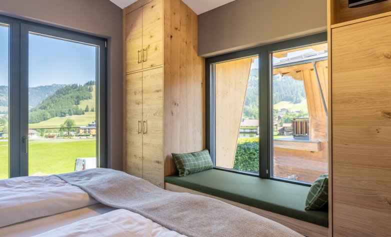 Helles Schlafzimmer mit großflächigen Fenstern und Blick auf die umliegende Berglandschaft. Warme Holzelemente, eine gemütliche Fensterbank mit grünen Kissen und natürliche Materialien schaffen eine harmonische Verbindung von Komfort und Natur. Ideal für erholsame Momente mit Panoramablick.