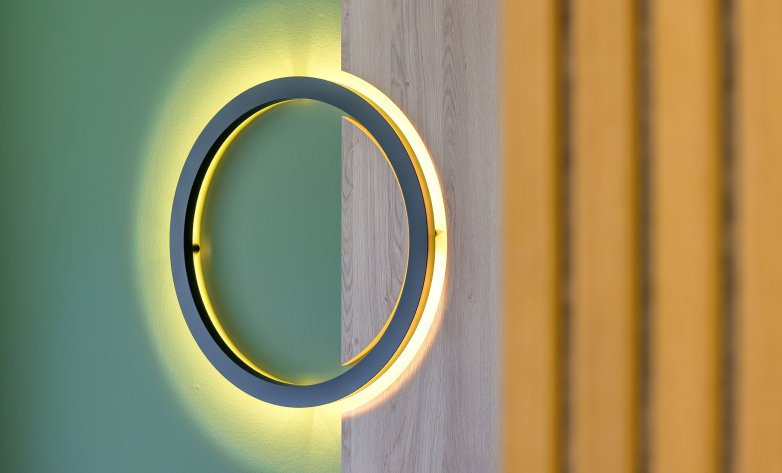 Detailaufnahme einer runden Wandleuchte mit indirekter LED-Beleuchtung. Der Lichtkreis ist halb in eine grün gestrichene Wand und halb in ein Holzelement integriert. Das warme Licht erzeugt einen weichen, atmosphärischen Schein und unterstreicht das moderne, hochwertige Raumdesign.