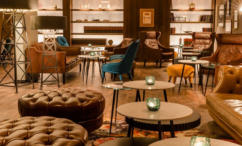 Stilvoll eingerichteter Lounge- und Barbereich mit gemütlichen Sitzmöbeln aus Leder und Stoff. Runde Tische mit kleinen Kerzen stehen zwischen gepolsterten Sesseln, Sofas und Hockern. Regale mit Flaschen und Gläsern sowie warme Lichtquellen im Hintergrund erzeugen eine einladende, entspannte Atmosphäre. Holzboden und dezente Farben unterstreichen das hochwertige, wohnliche Ambiente.