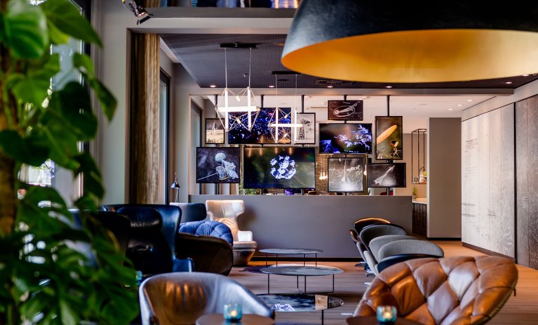 Eleganter Bar- und Loungebereich mit modernen Sitzmöbeln in dunklen Farben. Leder- und Stoffstühle sind um niedrige Tische angeordnet, darüber hängt eine große Design-Leuchte mit warmem Licht. Im Hintergrund befindet sich eine Bar mit beleuchteten Regalen, Gläsern und Flaschen. Pflanzen und Holzdetails lockern das Ambiente auf und sorgen für eine stilvolle, gemütliche Atmosphäre.