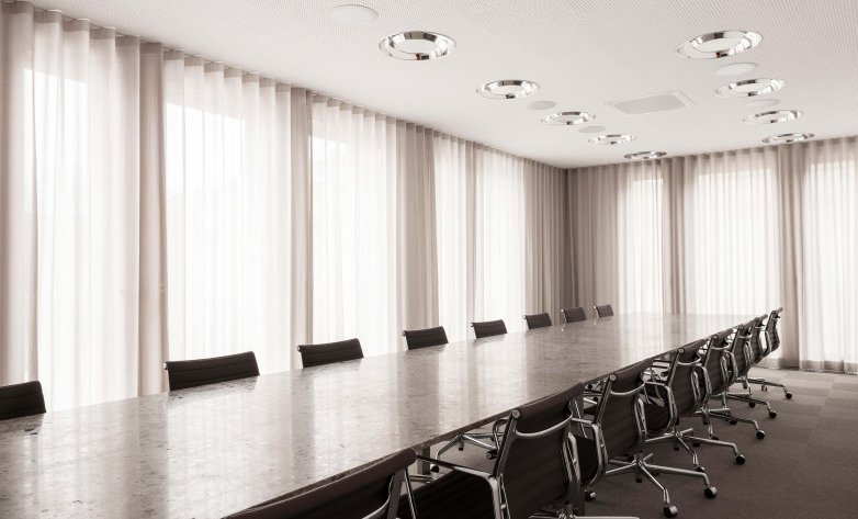 Großer, modern gestalteter Konferenzraum mit langem Besprechungstisch und mehreren schwarzen Drehstühlen. Bodentiefe, lichtdurchlässige Vorhänge sorgen für eine helle, gleichmäßige Ausleuchtung. Die klare Linienführung, dezente Farben und eingelassene Deckenleuchten schaffen eine ruhige, professionelle Arbeitsatmosphäre.
