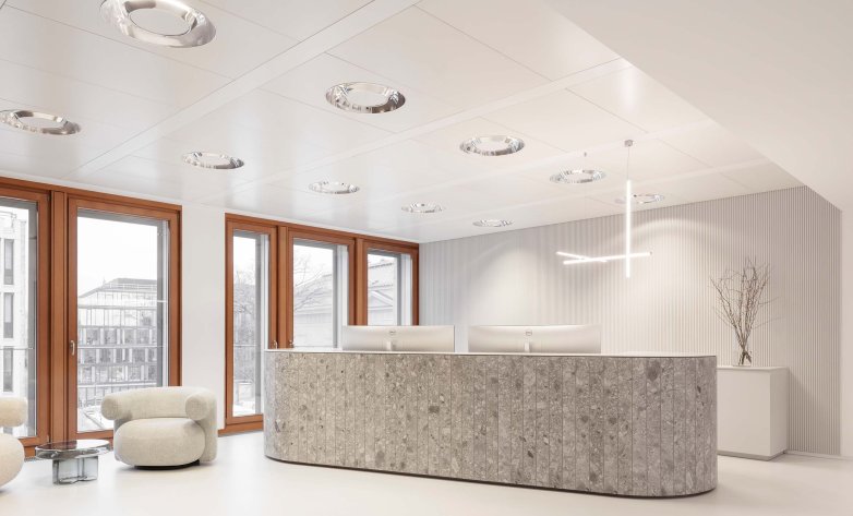 Moderner Empfangsbereich mit geschwungener, steinerner Empfangstheke und klarer, minimalistischer Gestaltung. Große Fenster mit Holzrahmen lassen viel Tageslicht herein. Dezente Farben, runde Einbauleuchten und eine filigrane Pendelleuchte schaffen eine ruhige, hochwertige Atmosphäre. Ein gemütlicher Sessel ergänzt den einladenden Eindruck.