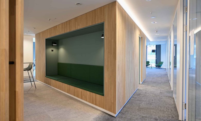 Moderner Büroflur mit grauem Teppich und heller Decke. Links befindet sich eine in Holzlamellen verkleidete Raumeinheit mit integrierter Sitznische in Grün, ausgestattet mit Polsterbank und Wandbeleuchtung. Rechts verläuft ein Gang mit Glastüren zu Büros. Am Ende des Flurs sind weitere Sitzmöbel und Tageslicht sichtbar.