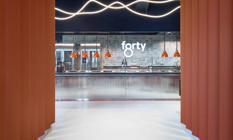Blick durch orangefarbene vertikale Lamellen auf eine moderne Bar mit glänzender Front und heller Arbeitsfläche. Dahinter eine geflieste Wand mit dem Schriftzug „forty“. Über der Theke hängen mehrere orange Pendelleuchten, darüber verlaufen geschwungene Lichtlinien an der dunklen Decke. Der Raum wirkt klar strukturiert und hochwertig gestaltet.