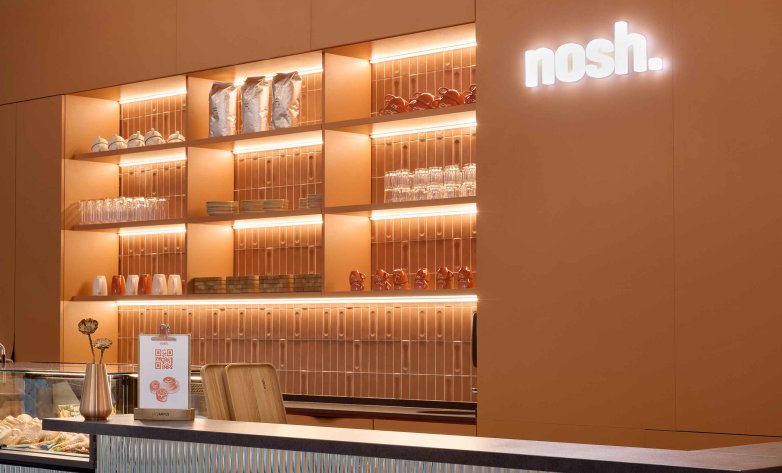 Modern gestalteter Thekenbereich in warmen Orangetönen mit hinterleuchteten Regalen. Darin stehen Gläser, Tassen, Schalen und dekorative Objekte. Rechts leuchtet der Schriftzug „nosh.“ an der Wand. Im Vordergrund befindet sich eine Theke mit kleiner Speisekarte, Tabletts und dekorativer Vase. Die Beleuchtung wirkt weich und einladend.
