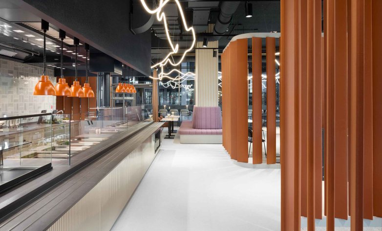 Moderner Gastronomieraum mit offener Theke und Glasvitrine links, darüber mehrere orange Pendelleuchten. Rechts strukturieren orangefarbene Lamellen den Raum. Im Hintergrund befindet sich eine rosafarbene Sitzbank mit Tischen. Geschwungene Lichtlinien an der Decke sorgen für eine dynamische, moderne Atmosphäre.