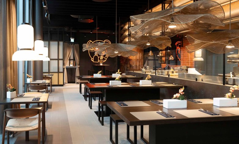 Modern eingerichteter Restaurantbereich mit langen Gemeinschaftstischen, dunklen Holzoberflächen und integrierter Showküche. Über den Tischen hängen große, organisch geformte Deckenelemente aus filigranem Netzmaterial. Bodentiefe Fenster, warme Beleuchtung und dezente Blumenarrangements schaffen eine einladende Atmosphäre. Das Design verbindet industrielle Elemente mit hochwertigen Materialien und moderner Gastlichkeit.