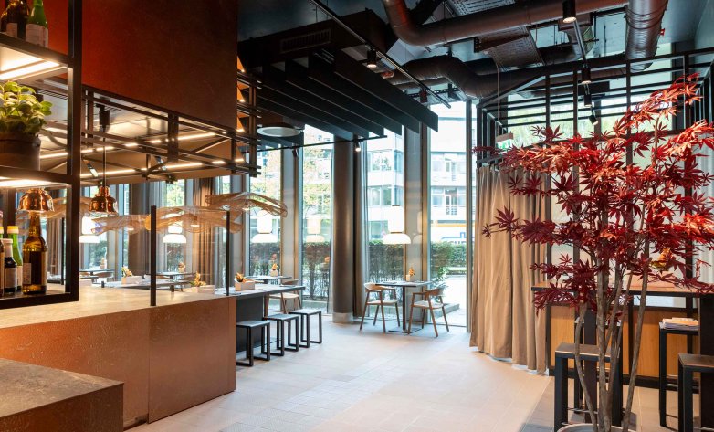 Moderner Gastronomiebereich mit offenem Raumkonzept, hohen Fensterfronten und großzügigem Tageslichteinfall. Dunkle Deckenstrukturen und hochwertige Materialien verleihen dem Raum einen industriell-eleganten Charakter. Ein dekorativer Baum mit roten Blättern setzt einen farblichen Akzent, während Sitzbereiche und eine offene Theke eine einladende Atmosphäre schaffen. Die Gestaltung verbindet Funktionalität, Ästhetik und moderne Gastlichkeit.