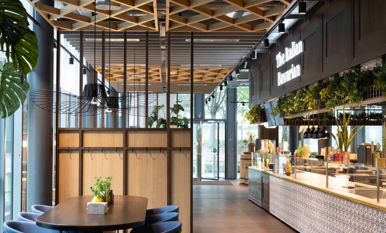 Modern gestalteter Restaurantbereich mit offenen Sitz- und Buffetzonen in einem hellen, einladenden Ambiente. Große Fensterfronten sorgen für viel Tageslicht, während Holzdecken, Pflanzen und hochwertige Materialien eine warme Atmosphäre schaffen. Eine moderne Speisenausgabe mit integrierter Begrünung bildet den zentralen Blickfang. Das Design verbindet Funktionalität, Natürlichkeit und zeitgemäße Gastronomiegestaltung.