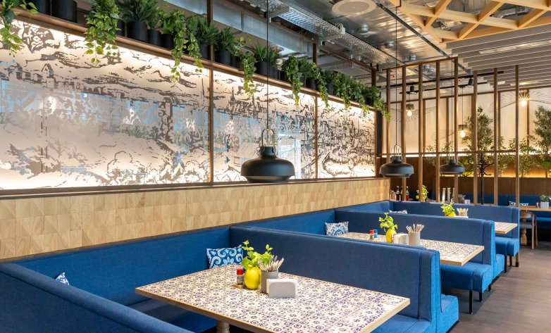 Moderner Restaurantbereich mit gemütlichen Sitznischen, blauen Polsterbänken und dekorativen Tischflächen. Eine großflächige, beleuchtete Wandgestaltung mit grafischen Motiven sowie integrierte Begrünung schaffen eine einladende Atmosphäre. Schwarze Pendelleuchten setzen gezielte Akzente, während Holz- und Metallelemente das moderne Raumkonzept ergänzen. Die Gestaltung verbindet Komfort, Funktionalität und zeitgemäßes Design.